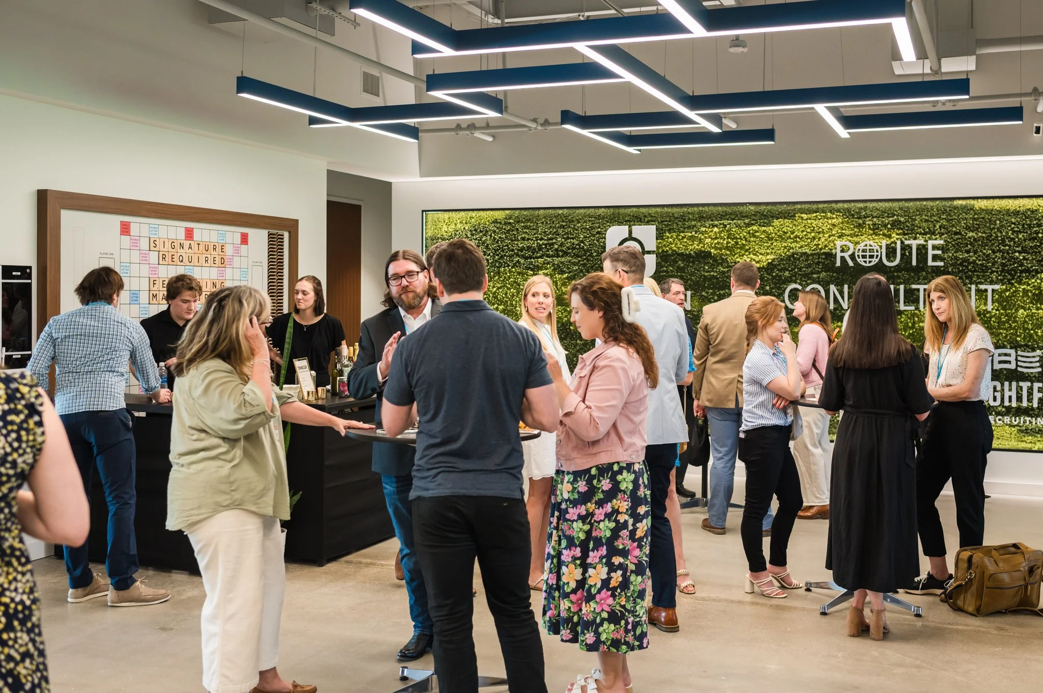 People networking and chatting at a modern conference or corporate event in an indoor space with a bright, industrial design. The background features a green wall with white text reading 'ROUTE CONSULTING' and a logo.