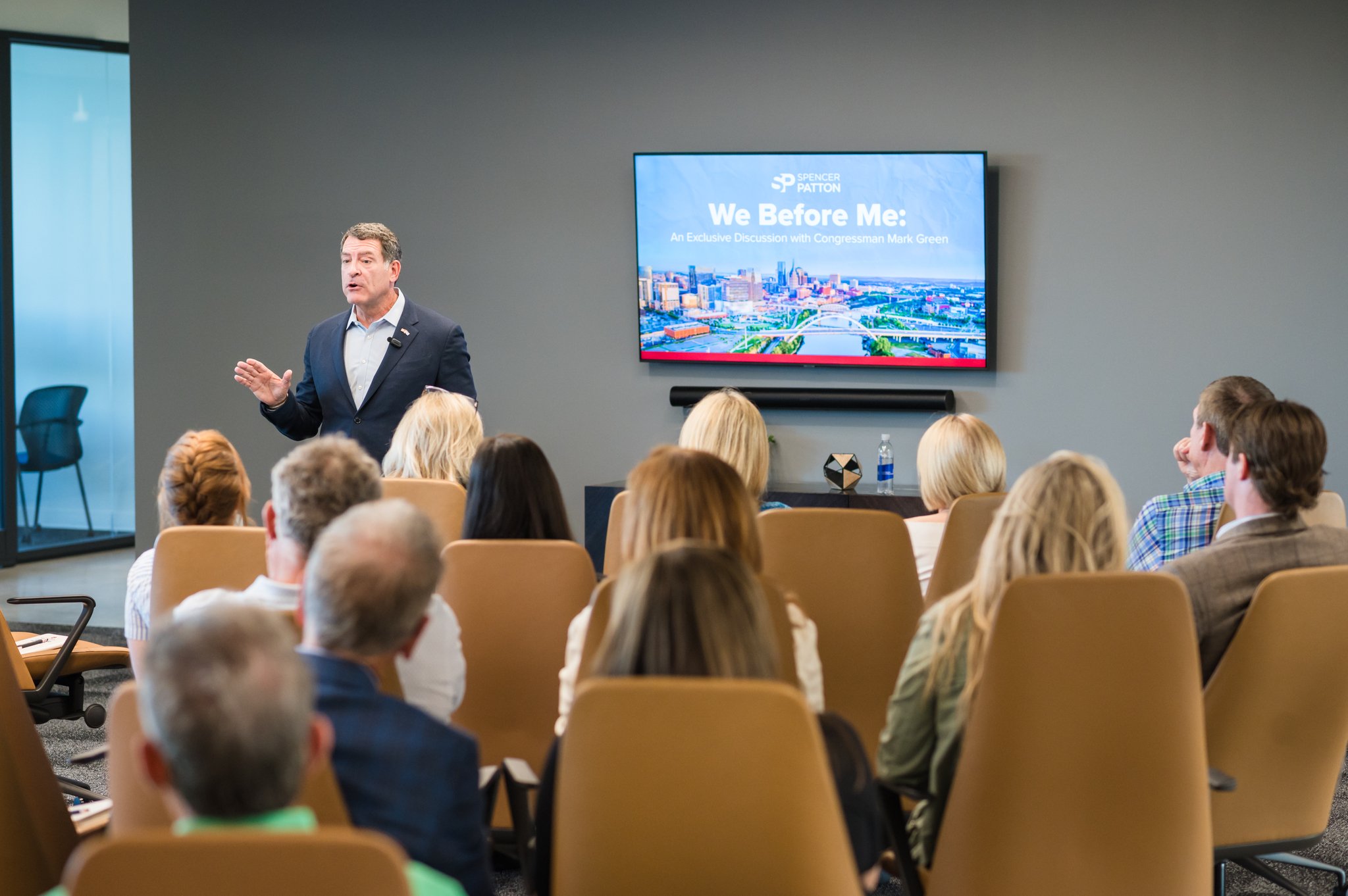 Man giving a presentation to an audience in a conference room with a screen displaying a slide titled 'We Before Me: An Exclusive Discussion with Congressman Mark Green'.