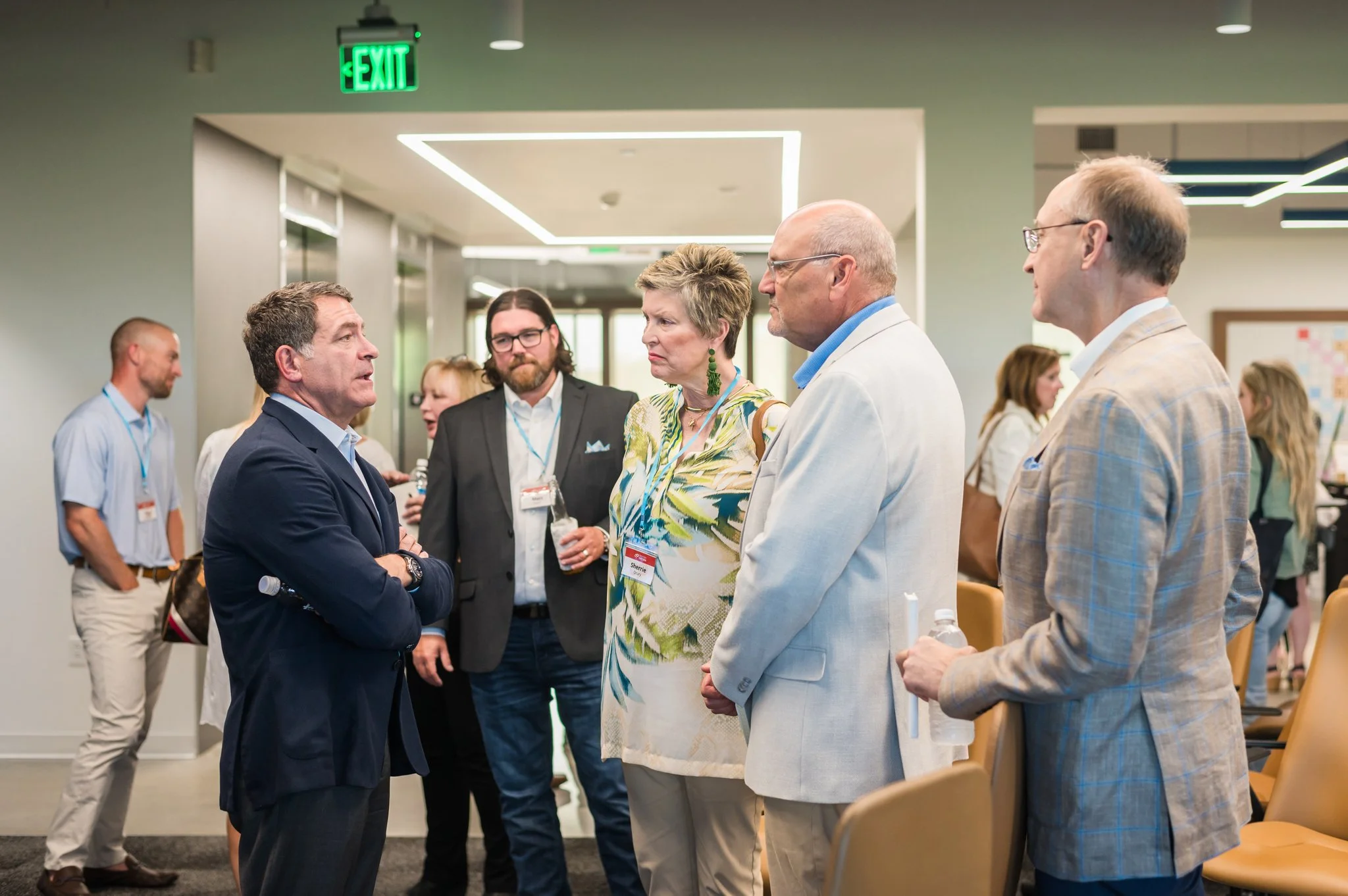 A group of professionals engaged in conversation at a conference or business event, with an exit sign visible on the wall behind them.