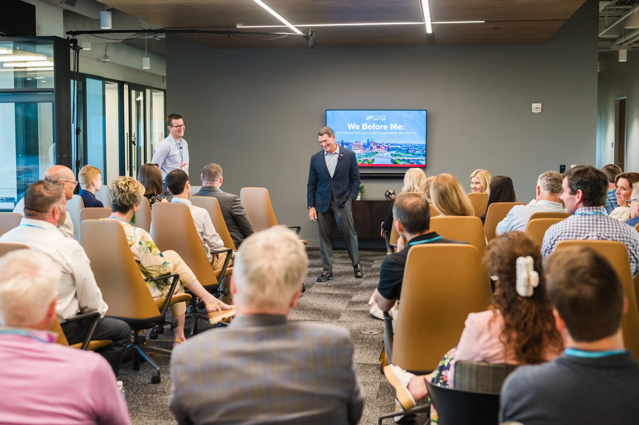 A professional conference room with a group of people seated in chairs, attentively listening to a speaker standing in front of a large screen. The speaker is dressed in a navy blue blazer and grey pants, smiling. Another person, wearing glasses and 