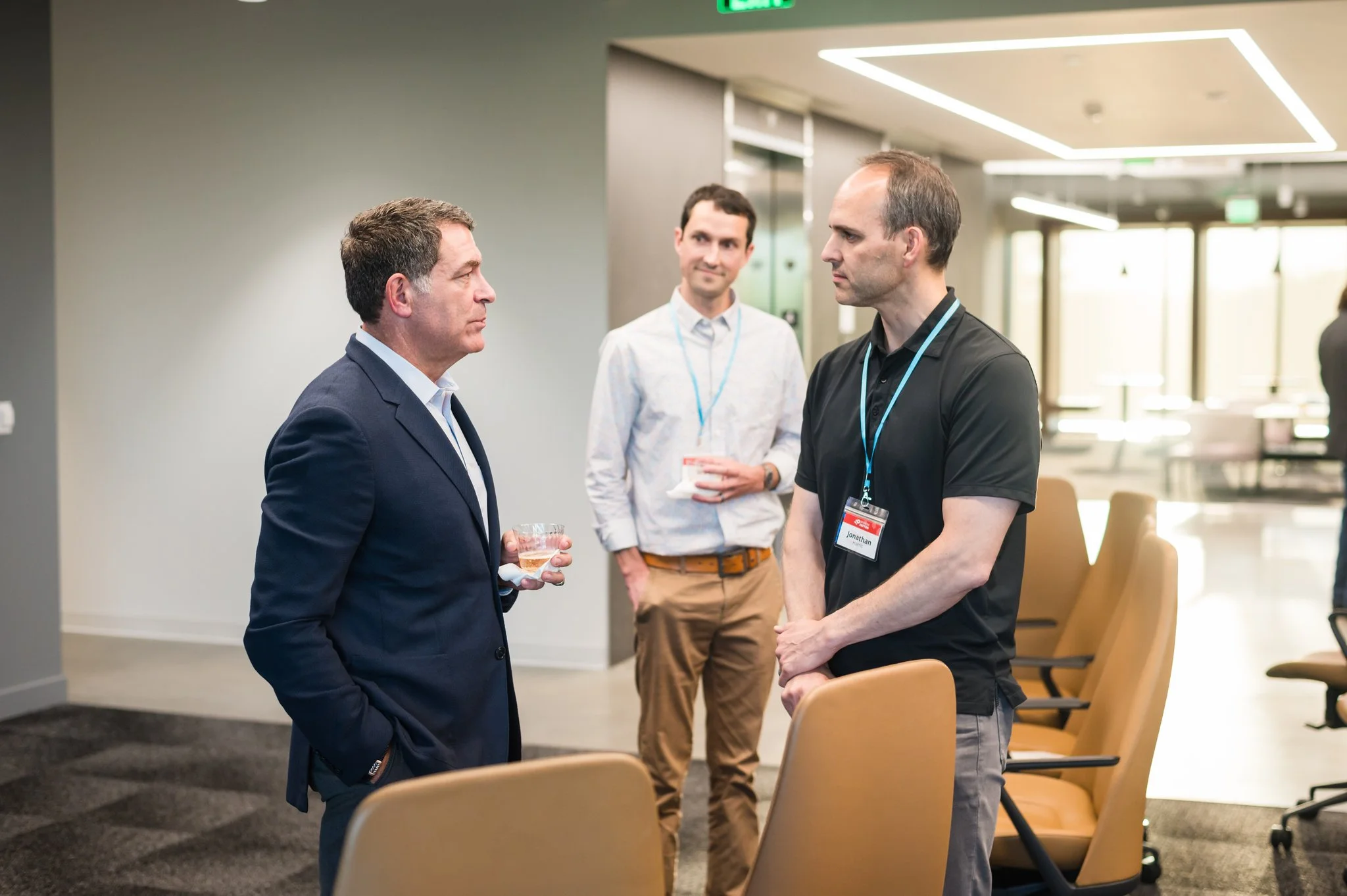 Three men having a conversation in a modern conference room, two of them wearing conference badges.