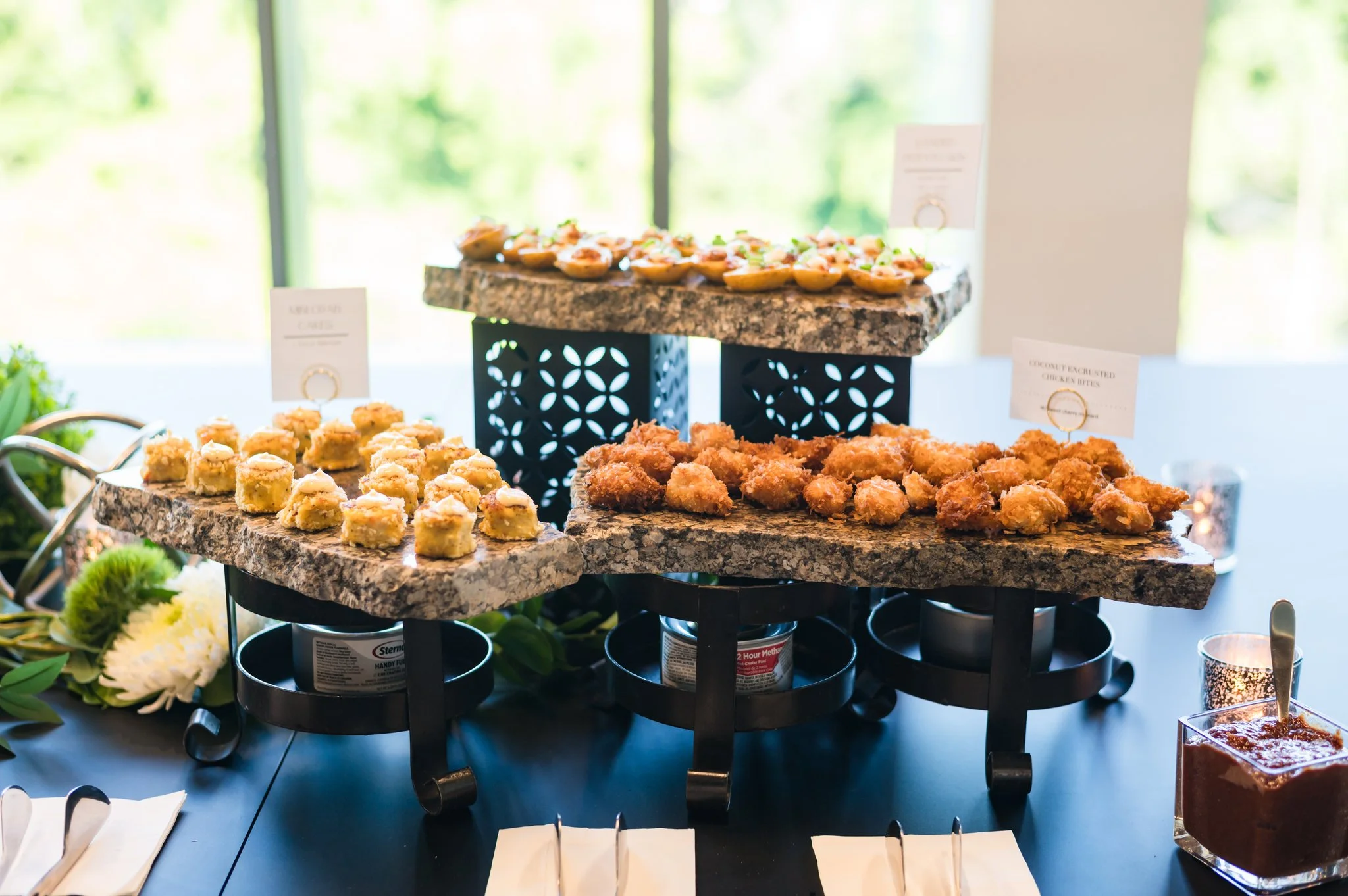 Assorted appetizers on three stone slabs, including small tartlets, fried chicken bites, and other finger foods, set on a black table with a dark tablecloth and decorative flowers nearby.