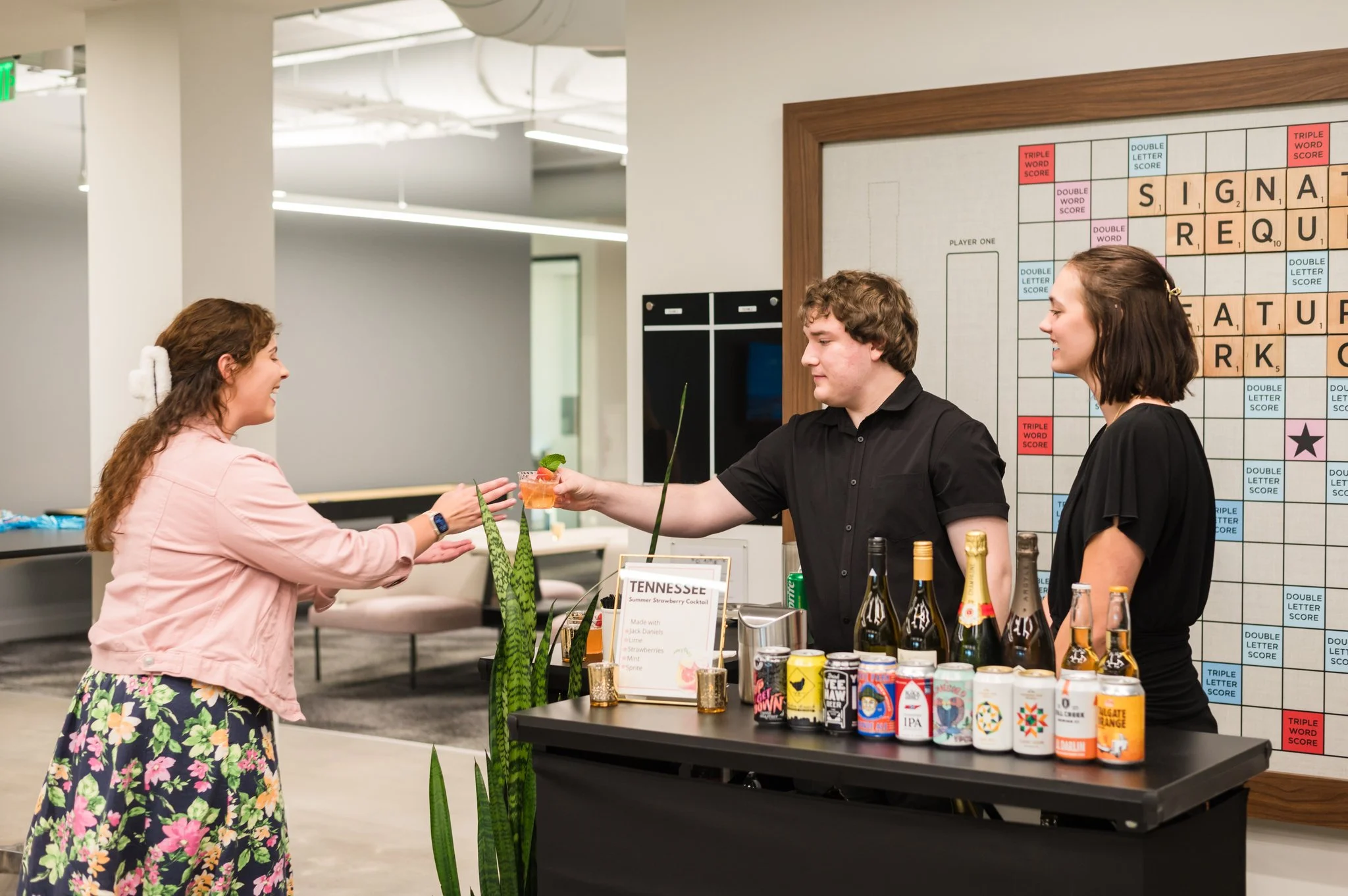 A woman in a pink jacket and floral skirt is receiving a cocktail from a bartender at a bar inside a modern office space. The bartender, wearing a black shirt, is handing over the drink, and a woman stands next to him. The bar has various bottles and