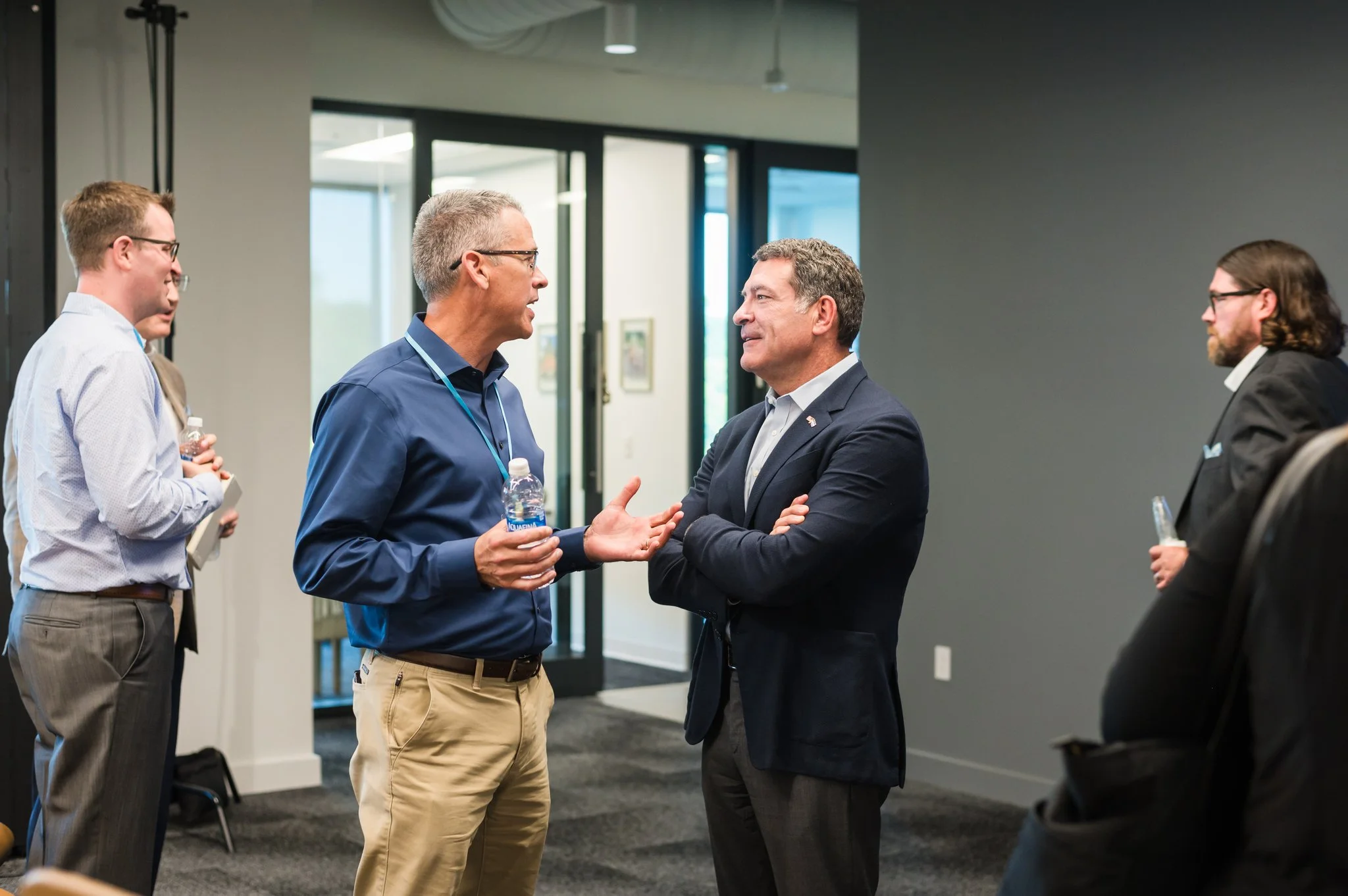 Two men engaged in conversation at a professional event, with other attendees nearby, in a modern office or conference setting.