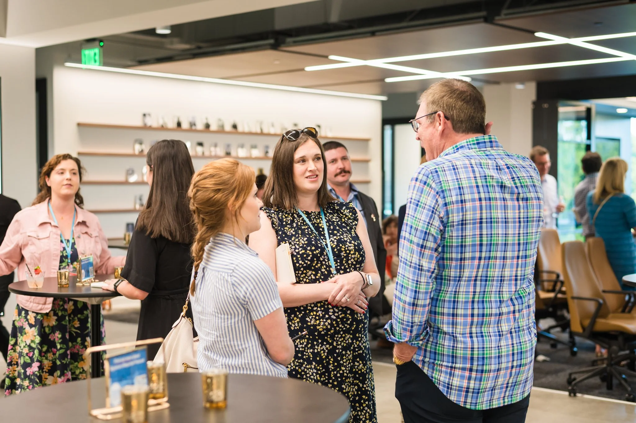 People socializing at a professional event in a modern, well-lit conference room with round tables, chairs, and decorative items on the shelves in the background.