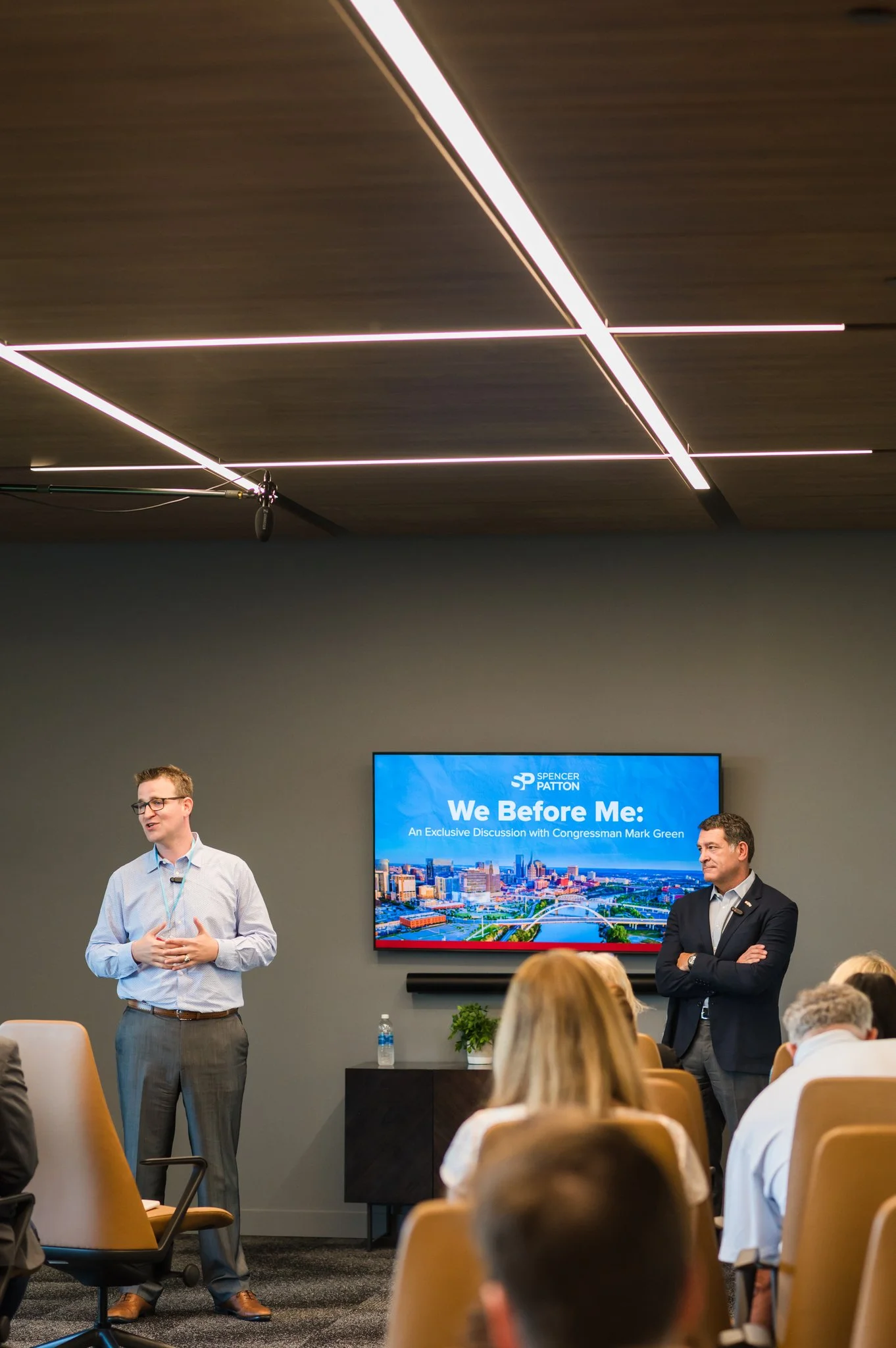A speaker is presenting at a conference with a slide titled 'We Before Me: An Exclusive Discussion with Congressman Mark Green' displayed on a screen behind him. The audience is seated, listening to the presentation in a modern conference room.