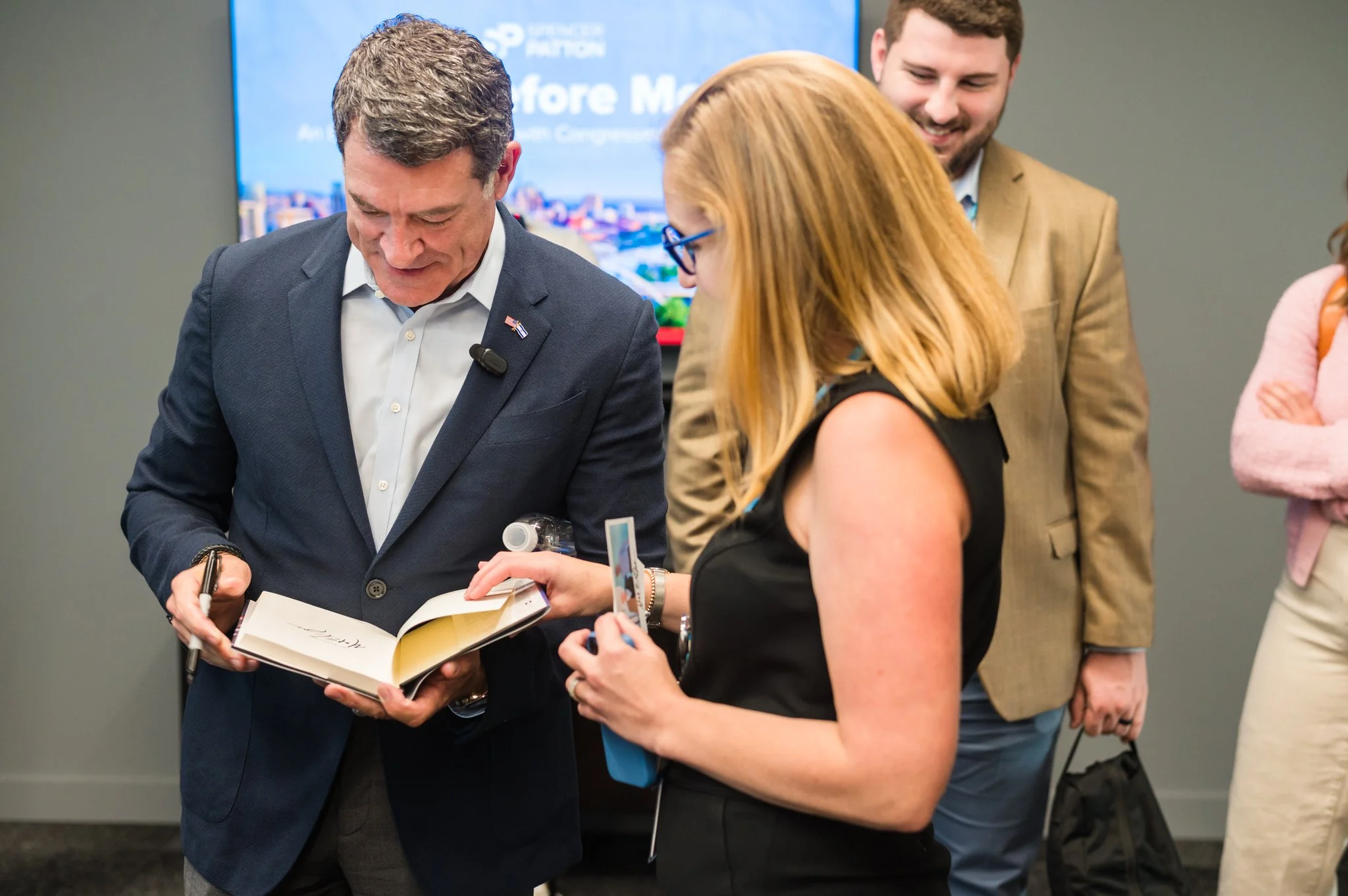 A man in a navy suit is signing a book for a woman with blonde hair and glasses, while another man in a tan suit watches and smiles in the background.
