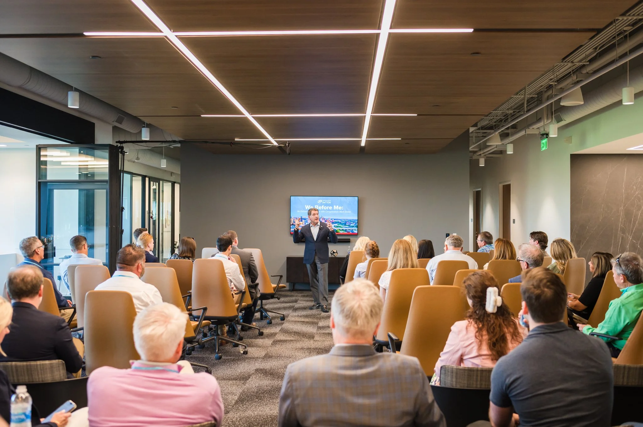 A man giving a presentation to an audience in a modern conference room with a TV screen displaying a slide titled 'We Before Me'.