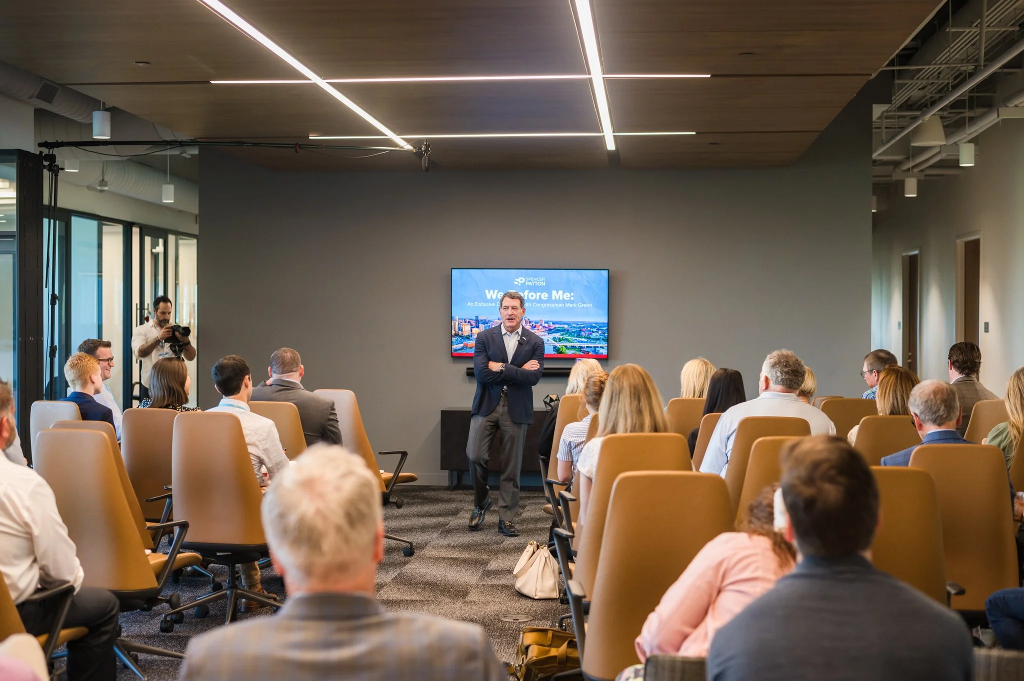 Man in suit giving a presentation to a seated audience in a modern conference room, with a large screen behind him displaying a slide.