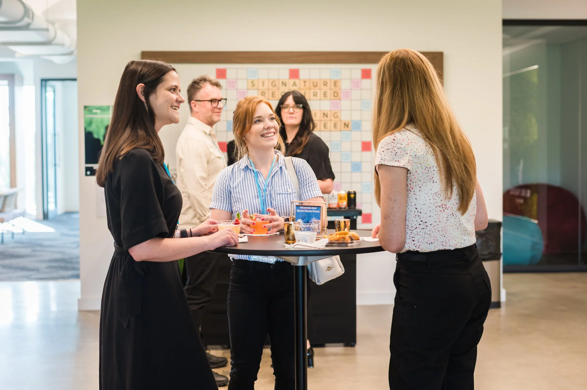 Group of five people talking and laughing at a social gathering in an indoor space with a writing board in the background.