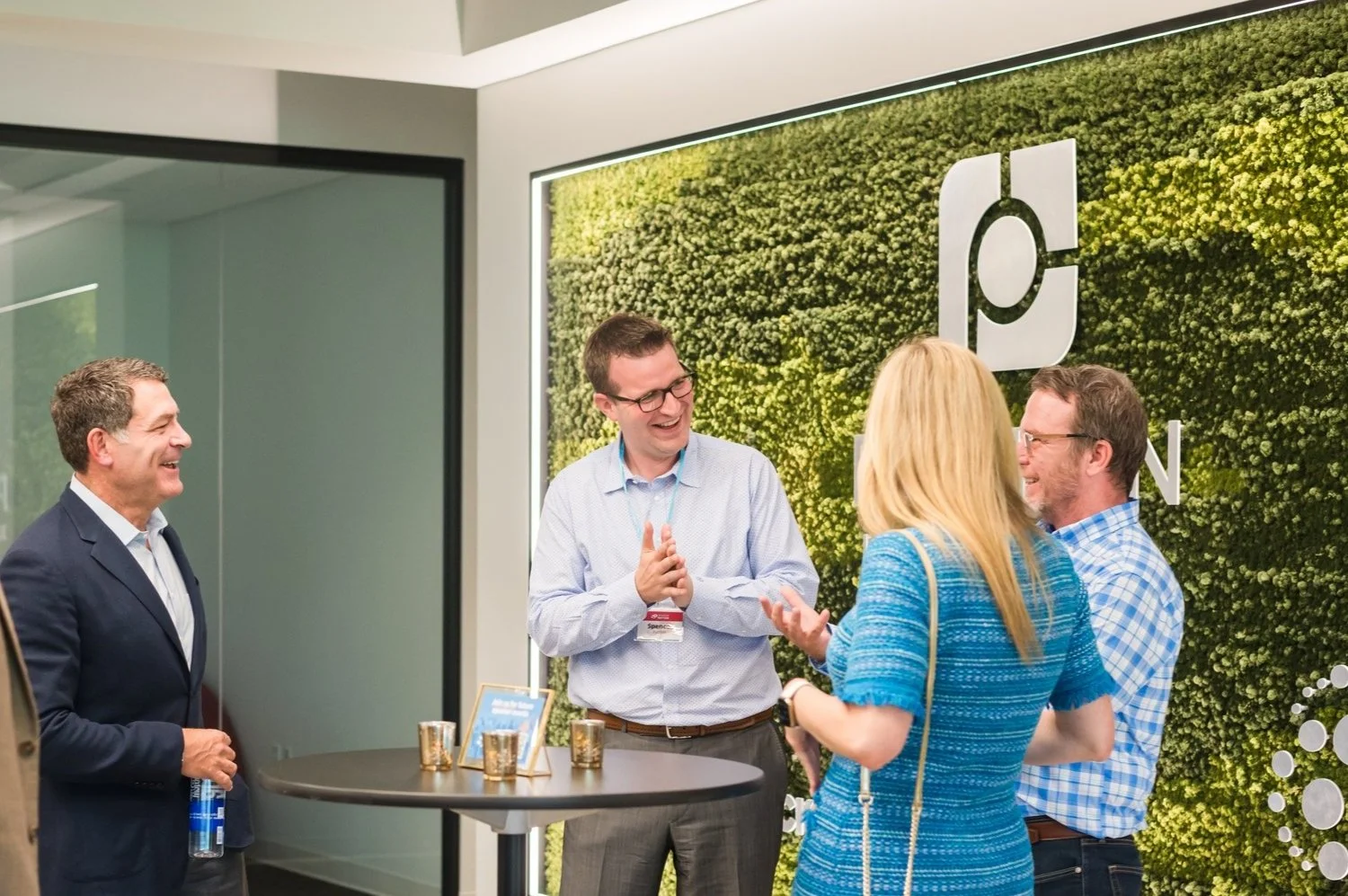 Group of five professionals in business casual attire engaging in conversation at a corporate event in front of a green wall with company logo.