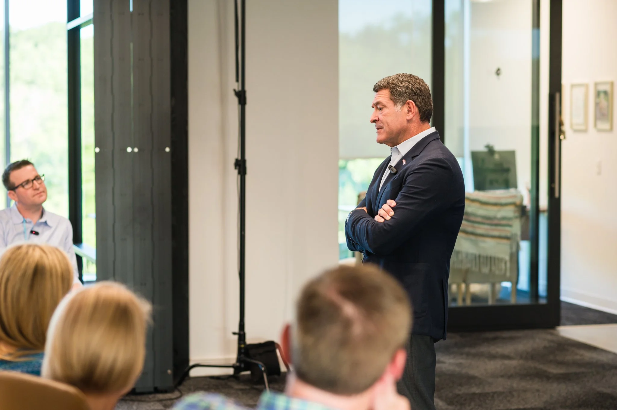 A man in a suit with arms crossed, giving a speech or presentation to a group of people in a room with large windows and artwork on the walls.