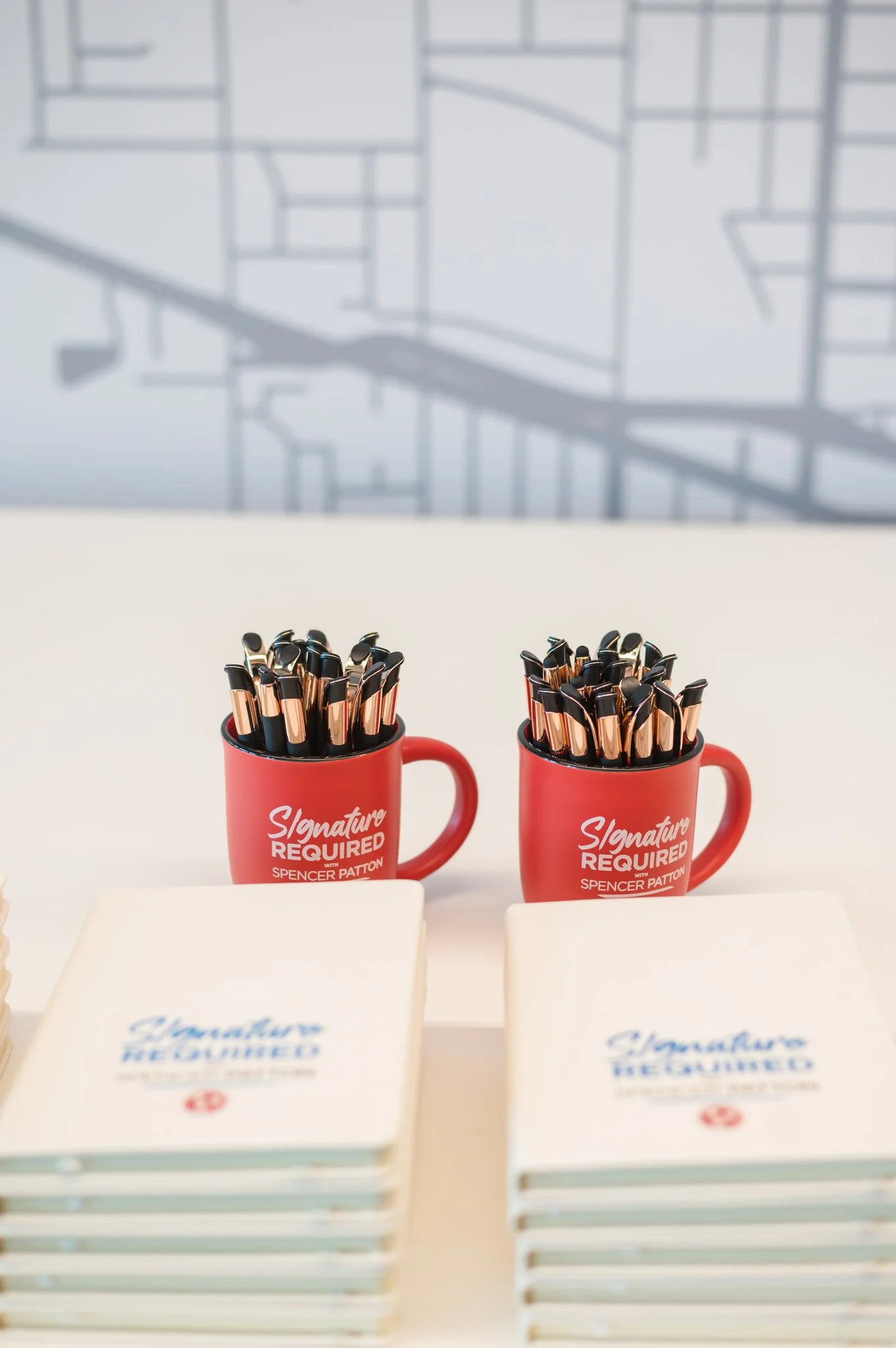 Two red mugs filled with black and gold pens on a white table, with stacks of signed copies of a book titled 'Signature Required' in front of each mug.