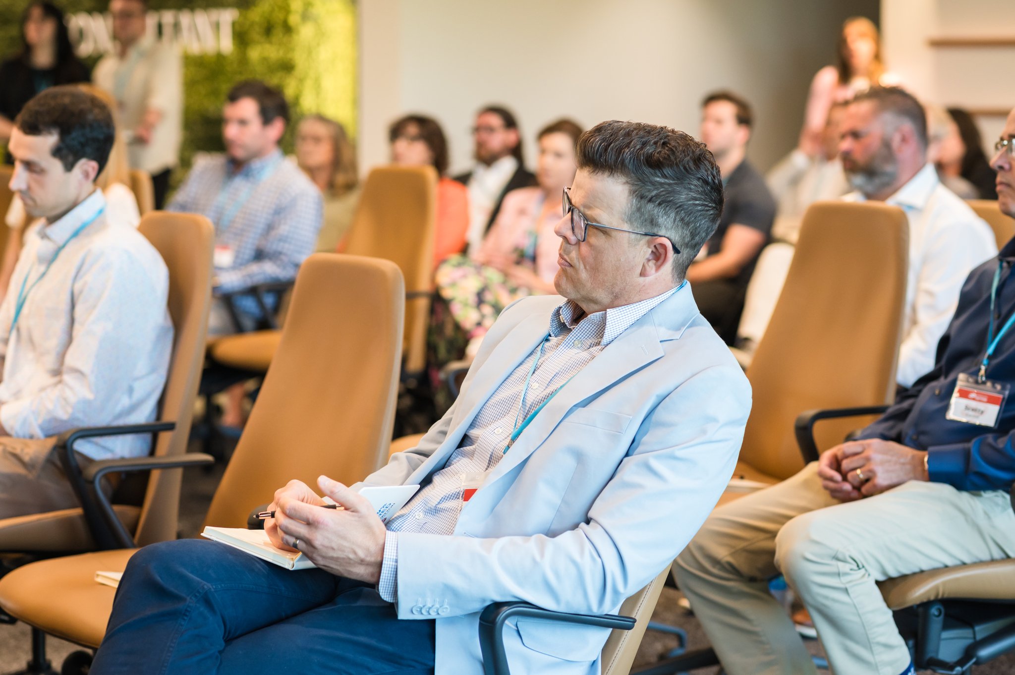 Business conference with attendees seated in rows, listening to a speaker, some taking notes.
