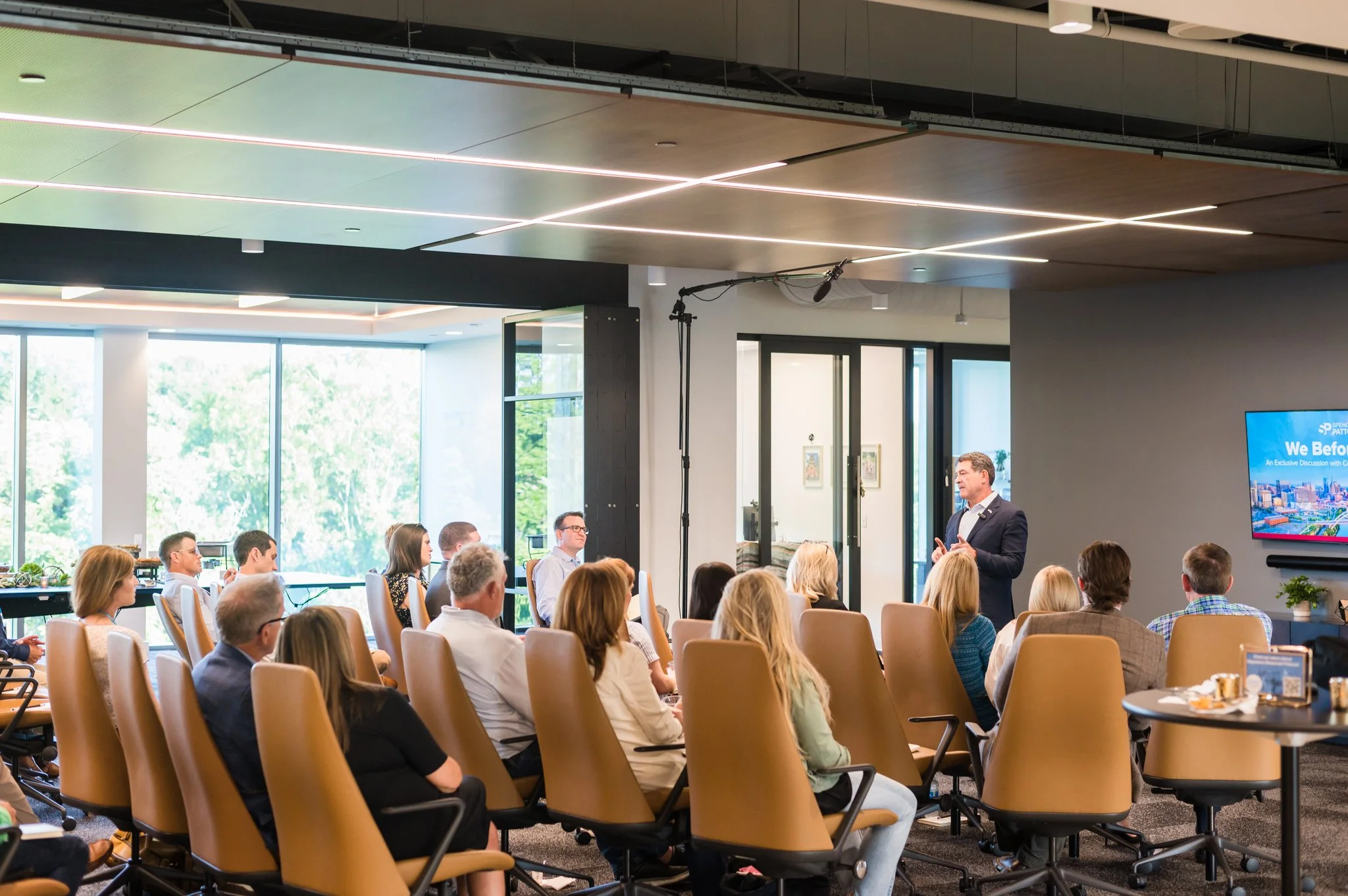 A businessman in a dark suit giving a presentation to a seated audience in a modern conference room with large windows showing green trees outside, and a large screen displaying a presentation slide.