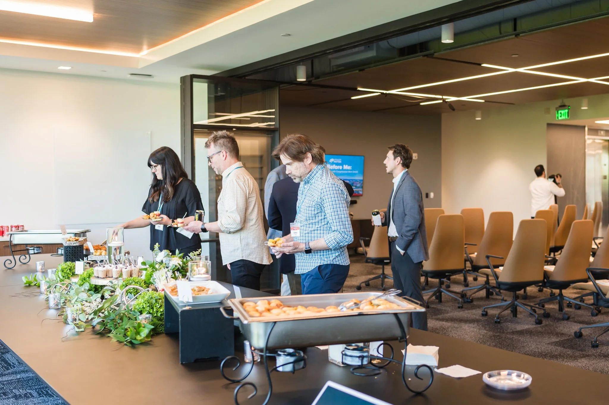 People in business casual attire at a catering buffet table in a conference room, selecting food and drinks during a break.