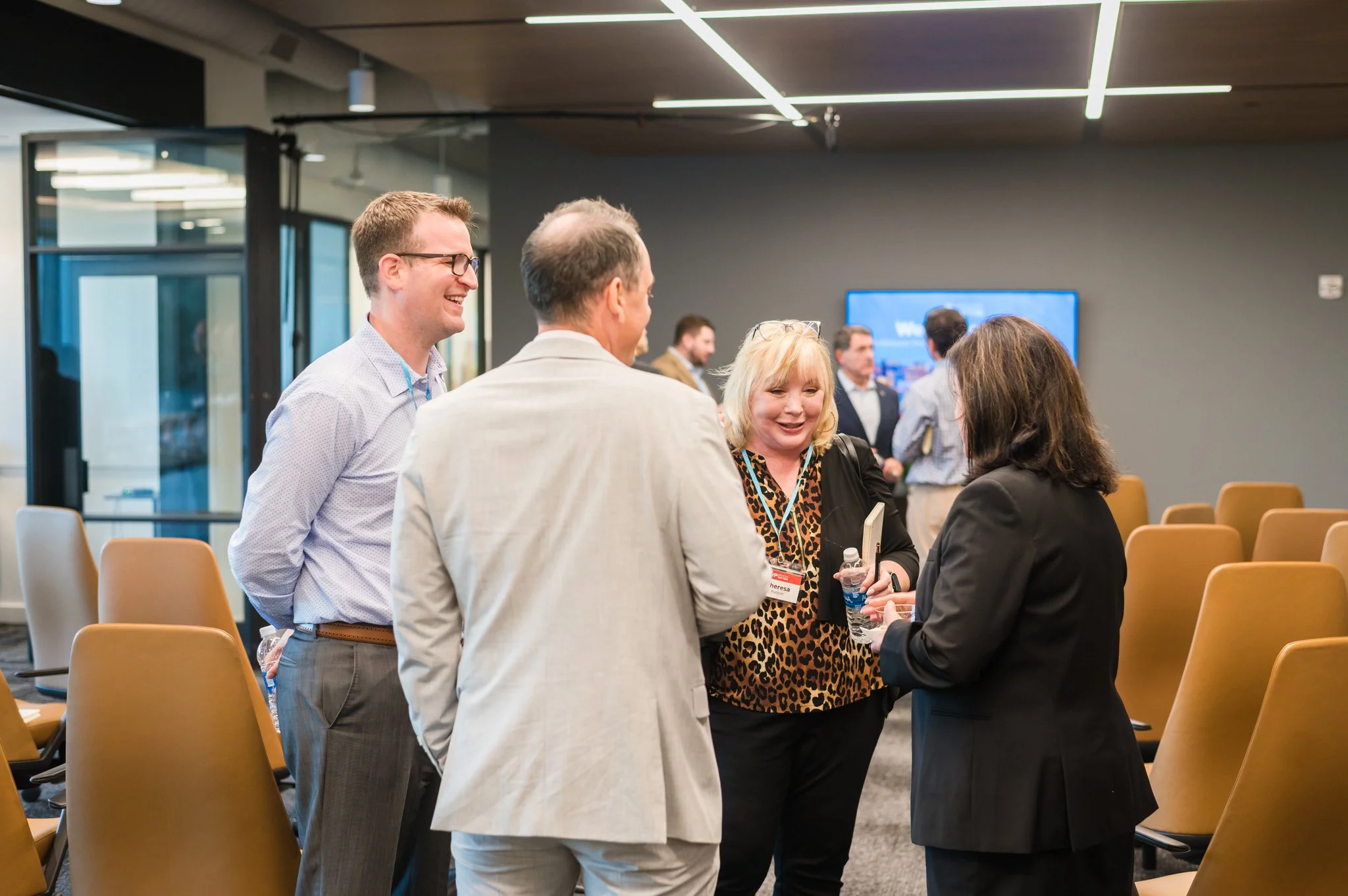 Group of four professionally dressed people talking and smiling at a conference or seminar in a modern room with yellow chairs and a large digital screen.
