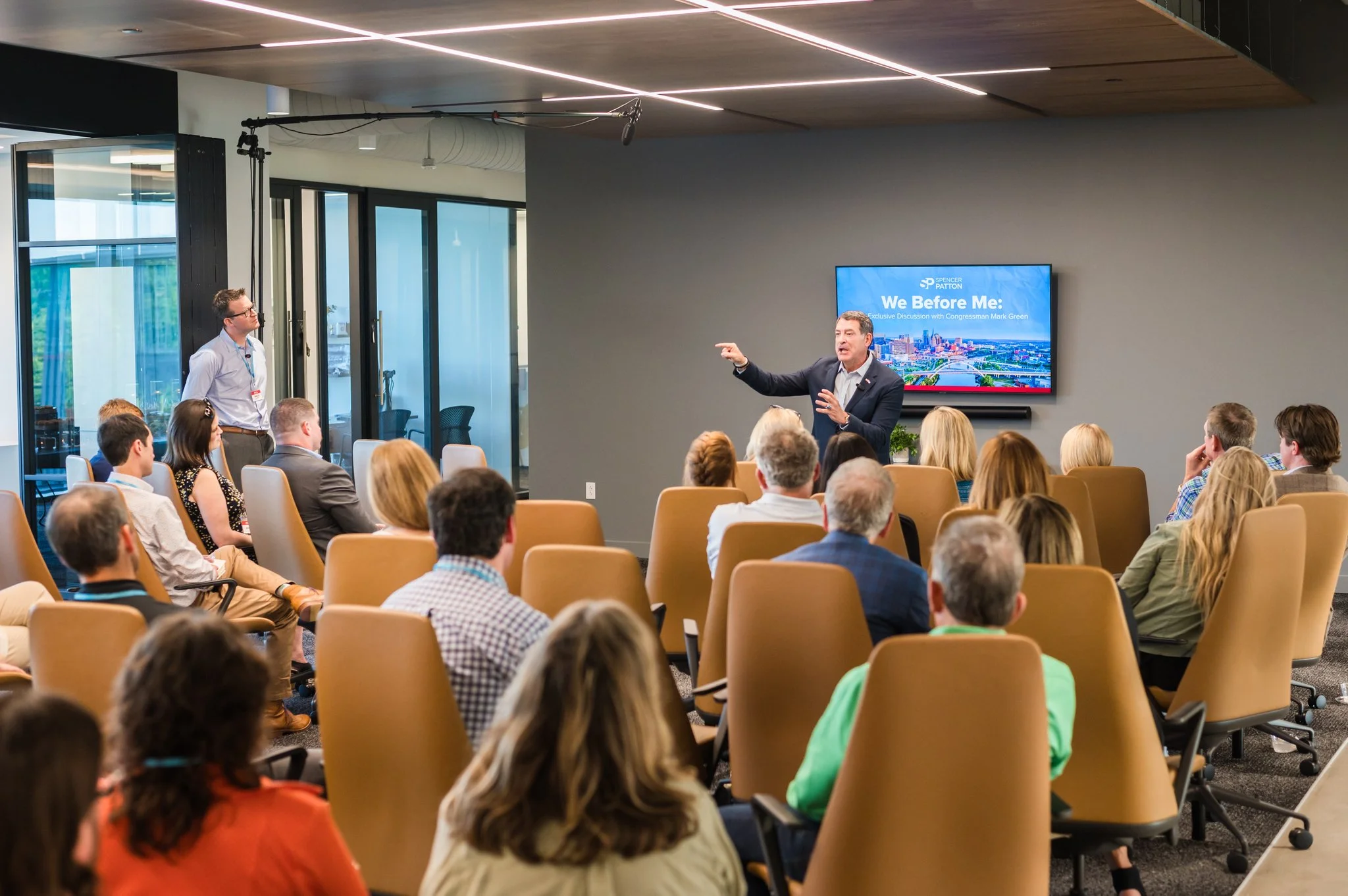 A man in a black blazer and white shirt giving a presentation, pointing towards a large screen that reads 'We Before Me:'. He is speaking to an audience seated in a modern conference room with tan chairs, and a woman stands near the back. The room ha