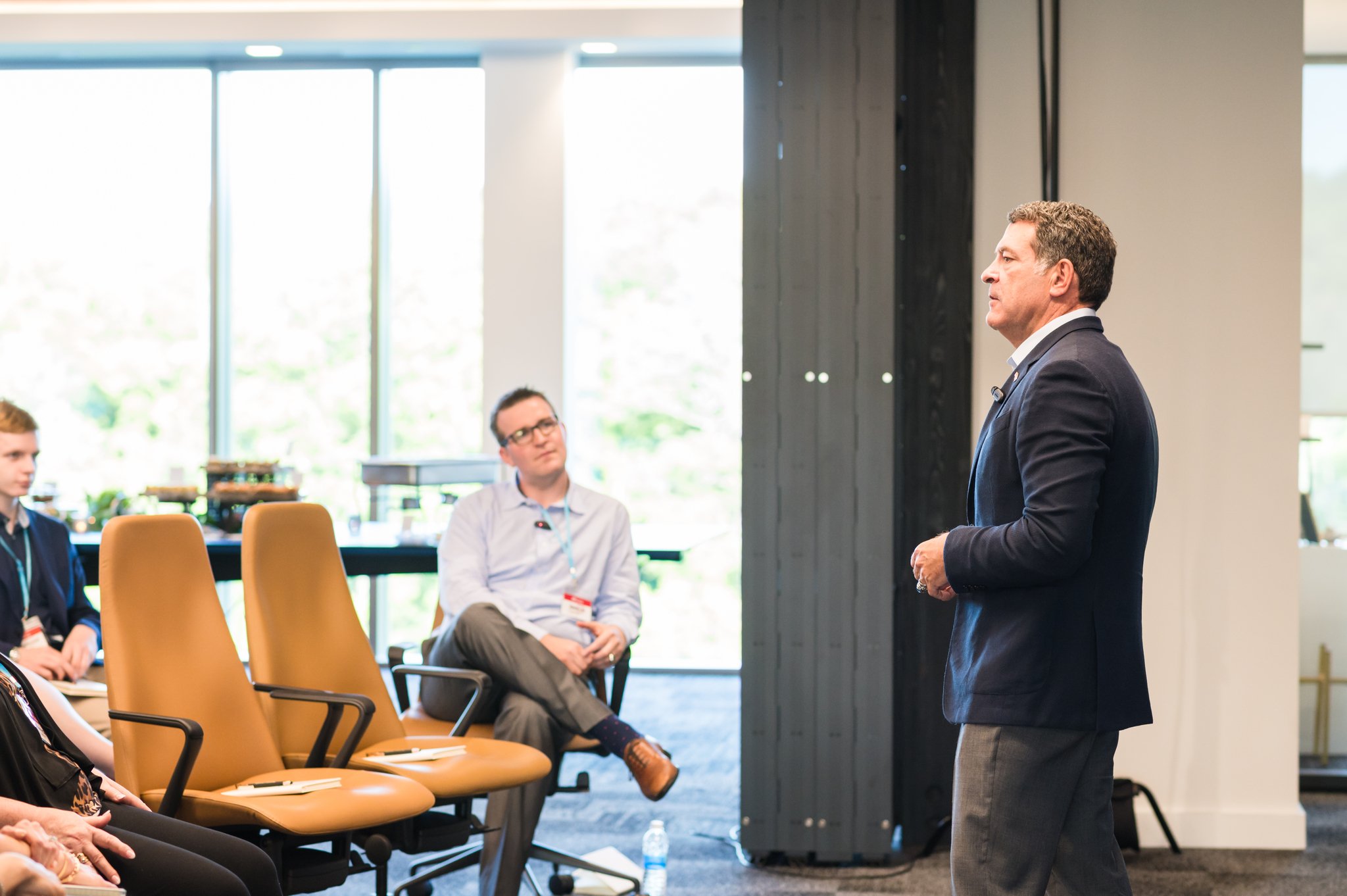 A man in a dark blazer giving a presentation in a conference room with seated attendees listening.