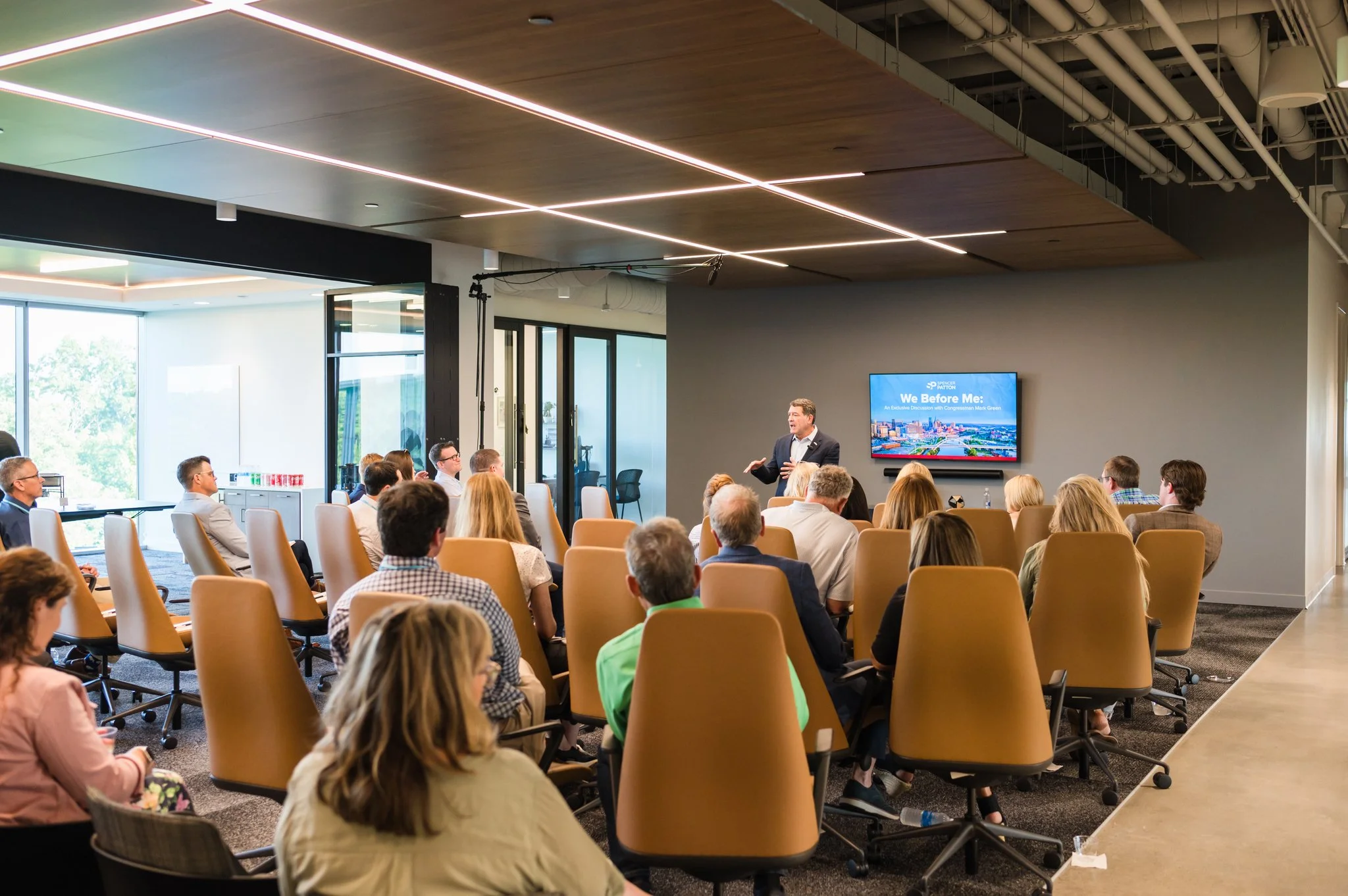 A man in a suit giving a presentation to an audience in a modern conference room with a large screen displaying the title 'We Before Me: An Exclusive Discussion with Congressman Mark Green'.