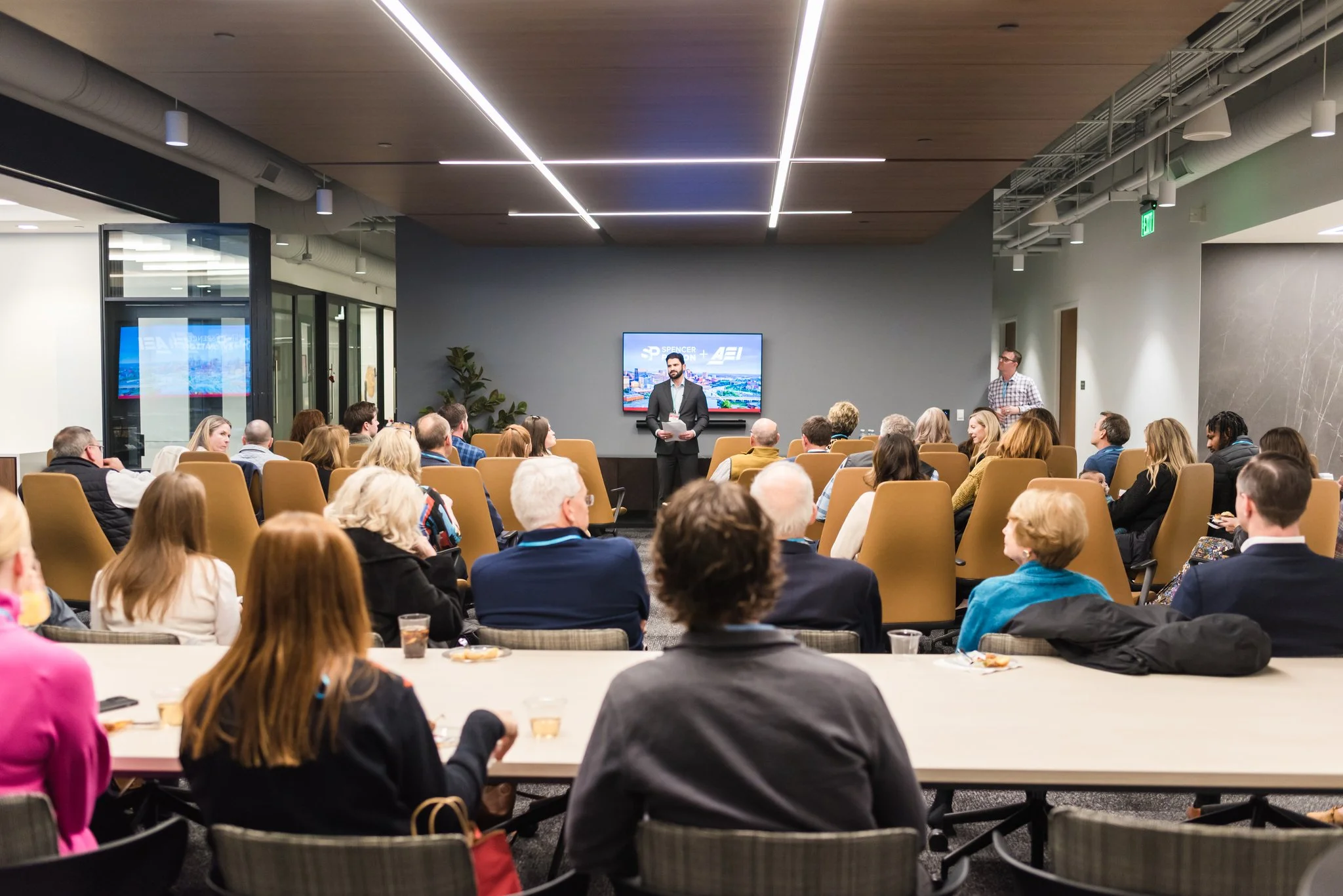 A group of people attending a formal presentation in a conference room. The speaker is standing in front of a screen, addressing the audience. The room has modern decor with wood accents and is filled with seated attendees.