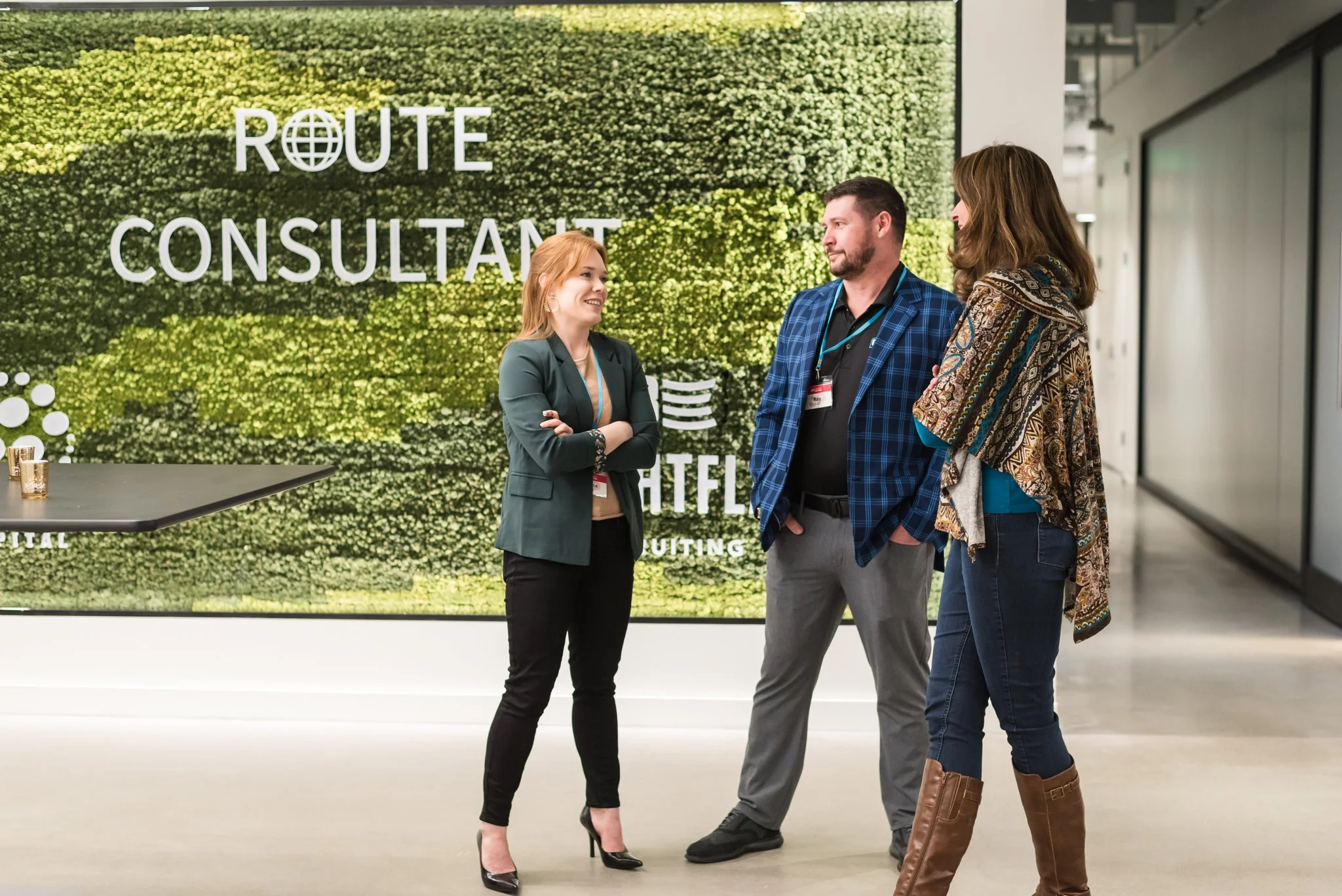 Three people standing and talking in front of a digital display showing 'ROUTE CONSULTANT' and the Netflix logo, inside a modern office or conference center.