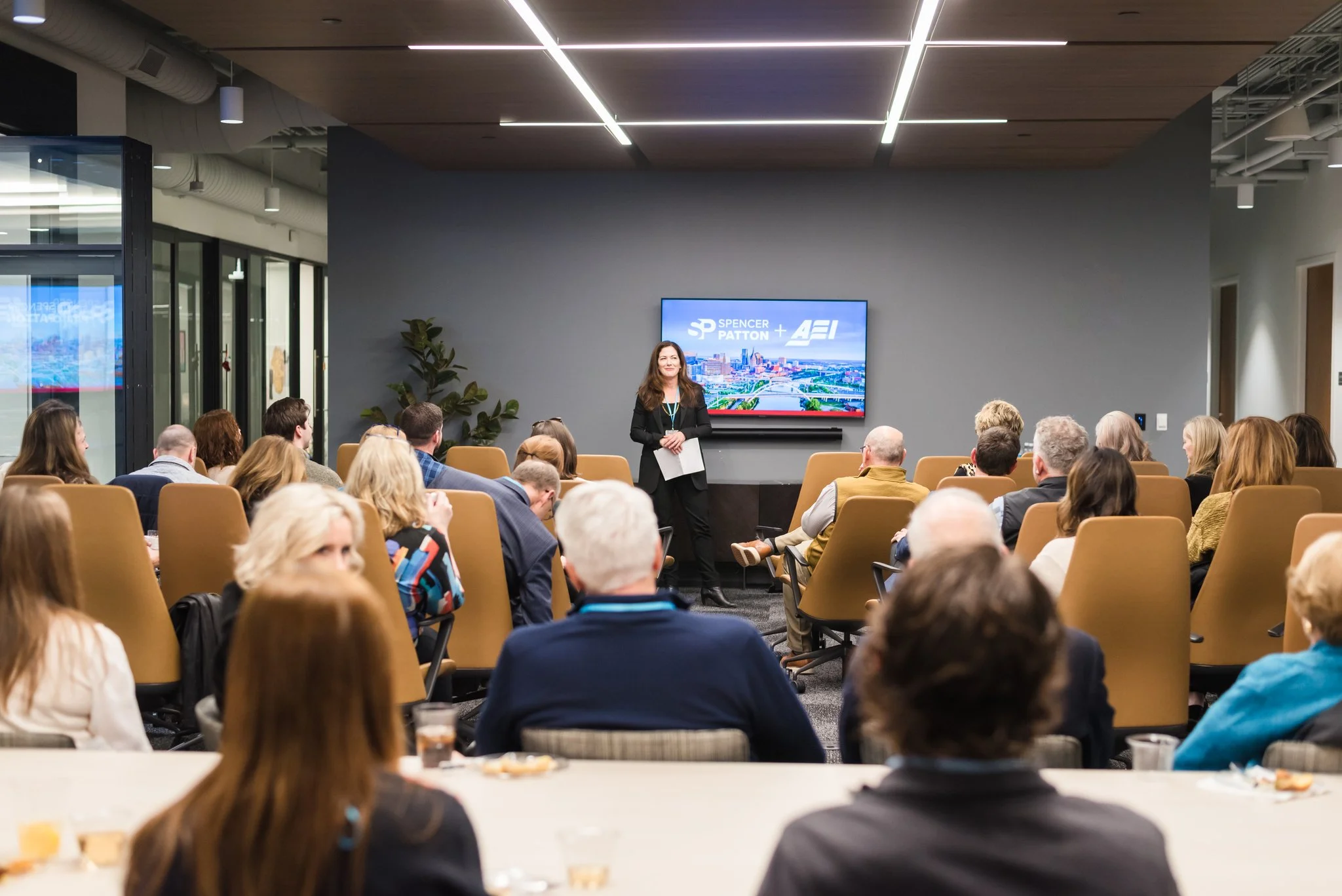 A woman in black standing in front of a group of people during a presentation in a modern conference room with a large screen displaying 'Spencer Patton + Afi'.