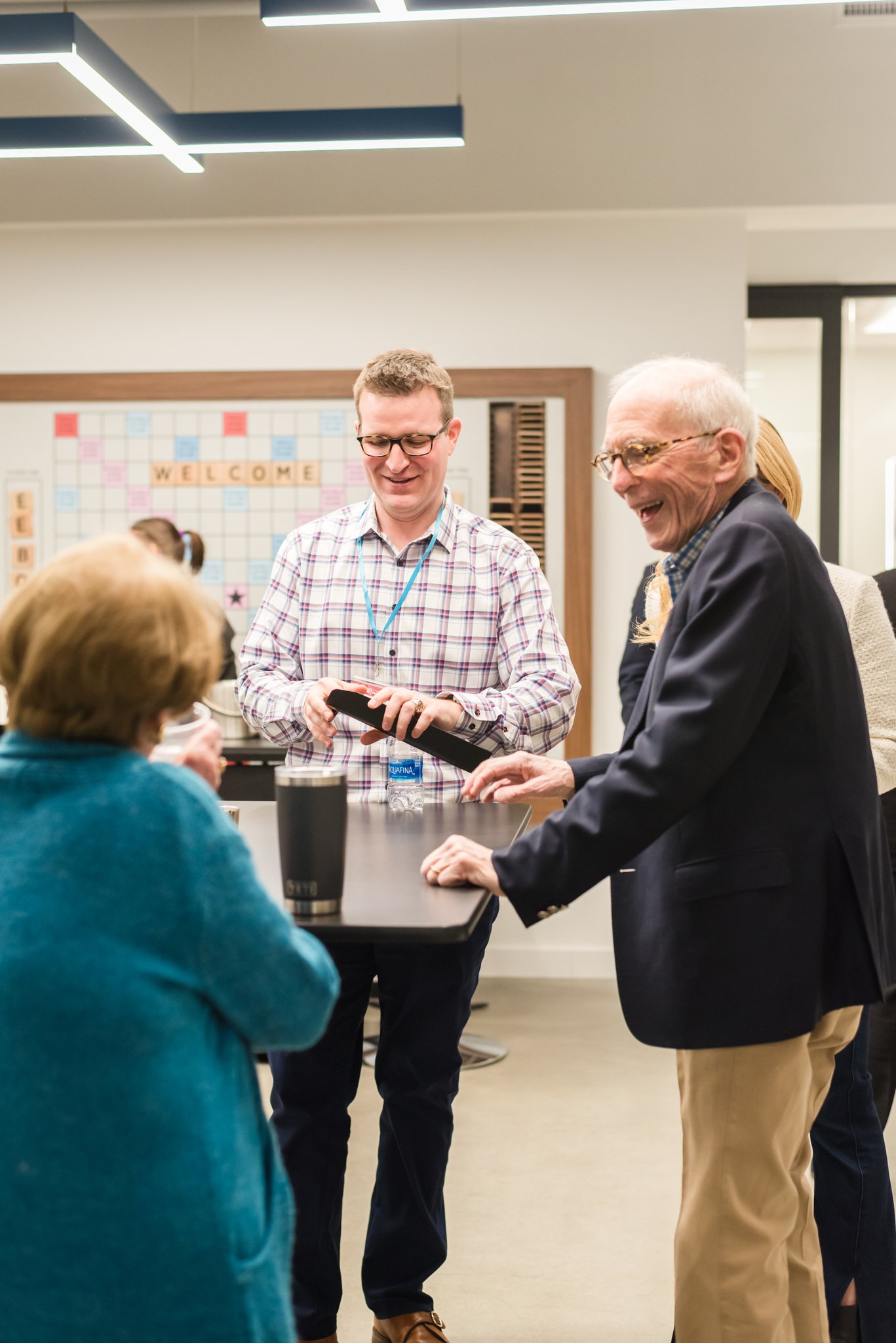 Four people, two women seated and two men standing, engaged in conversation and smiling in an office setting. One man is showing something on his phone.