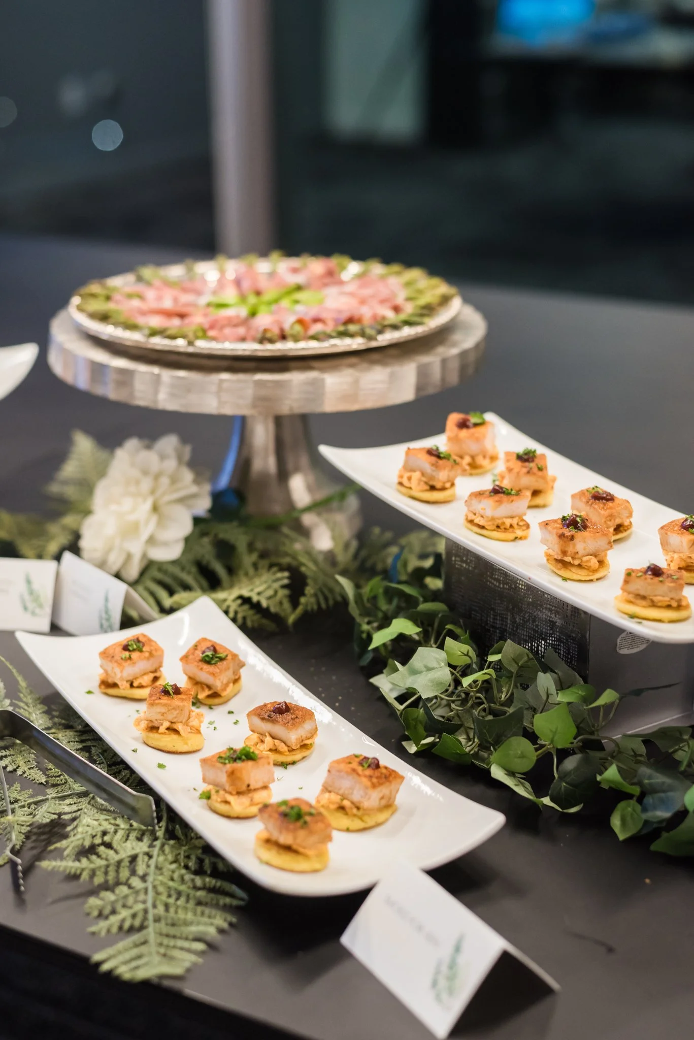 A display of assorted finger foods on white serving plates at a catering event, with a floral arrangement and greenery for decoration.