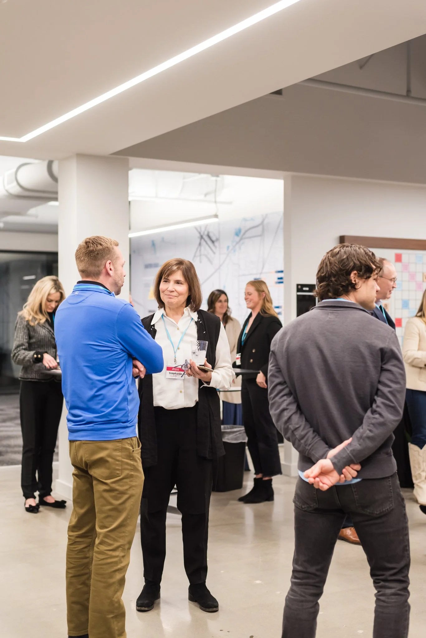 People socializing at a professional event in an office setting, with a woman holding a glass and phone, engaging in conversation with a man in a blue jacket.