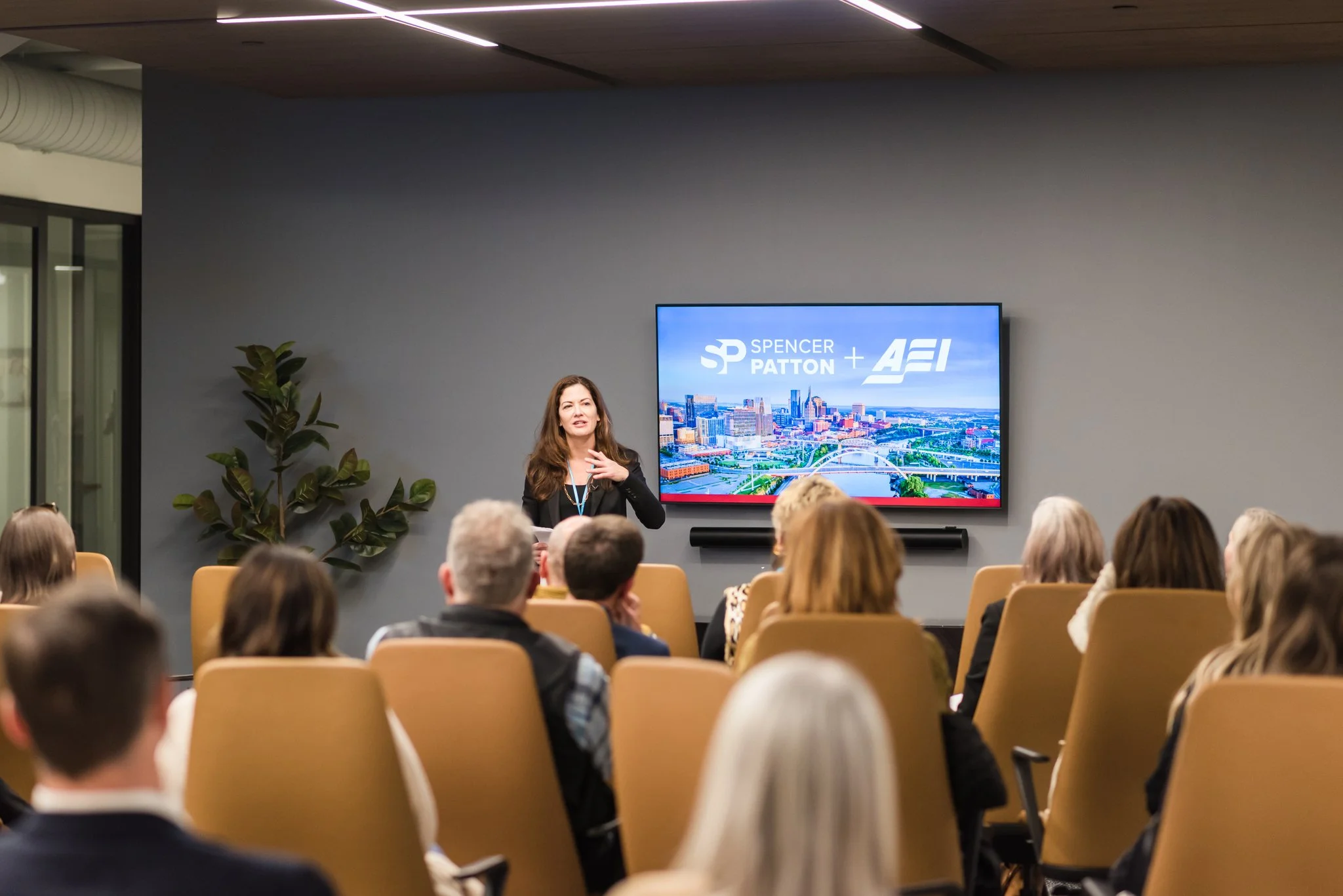 A woman presents to an audience in a conference room with a large screen behind her displaying the logos of Spencer Patton and AEI, and a cityscape image.