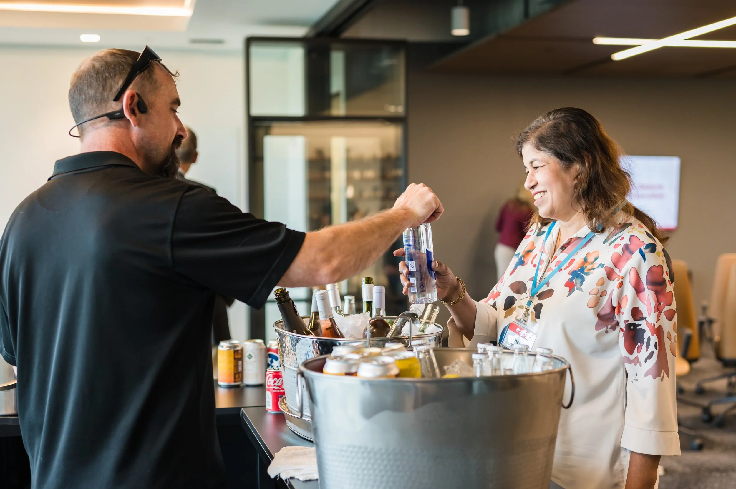A man in a black shirt and sunglasses handing a bottle of water to a smiling woman in a floral blouse at a beverage station with bottles and cans of drinks.