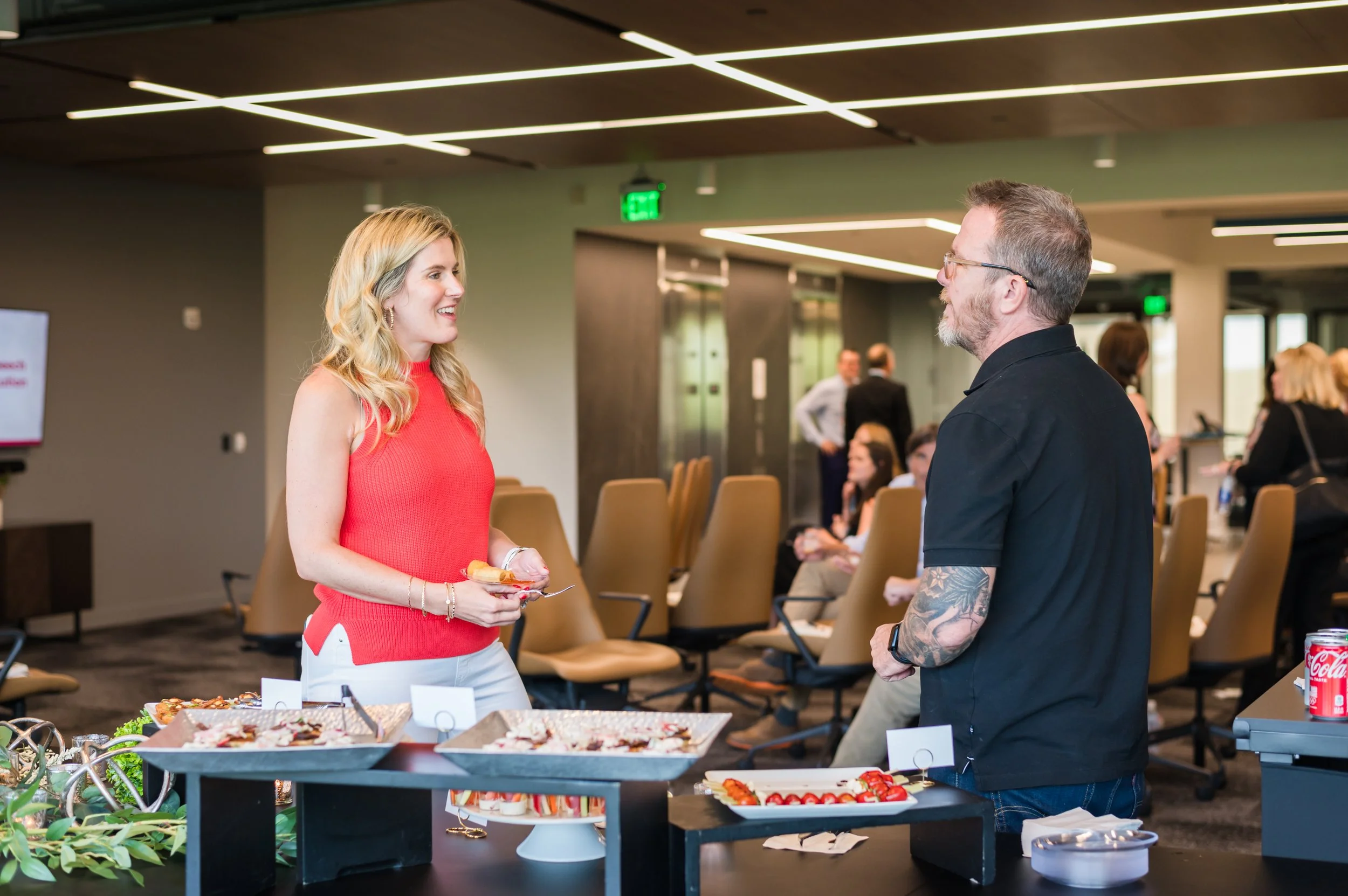 Two people exchanging smiles at a catering table during a professional event, with additional attendees in the background.