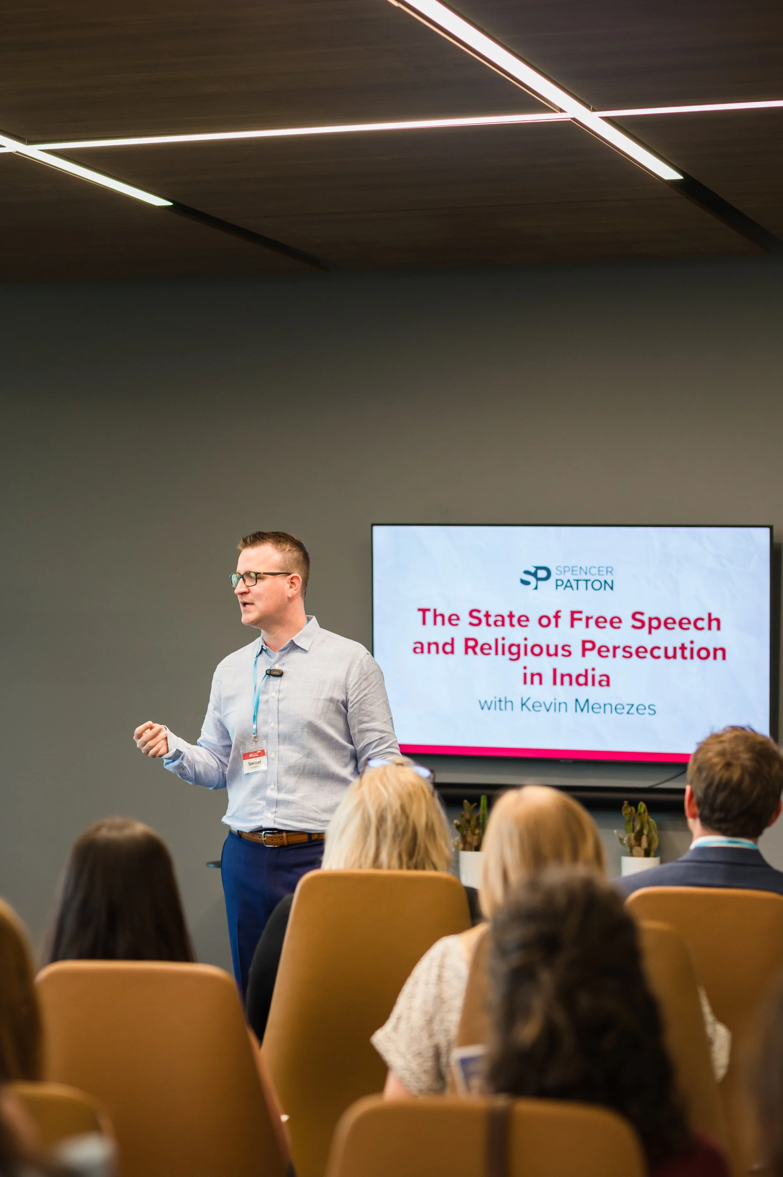 A man is giving a presentation in front of an audience in a conference room. The presentation slide on the screen behind him reads 'The State of Free Speech and Religious Persecution in India' by Kevin Menezes.