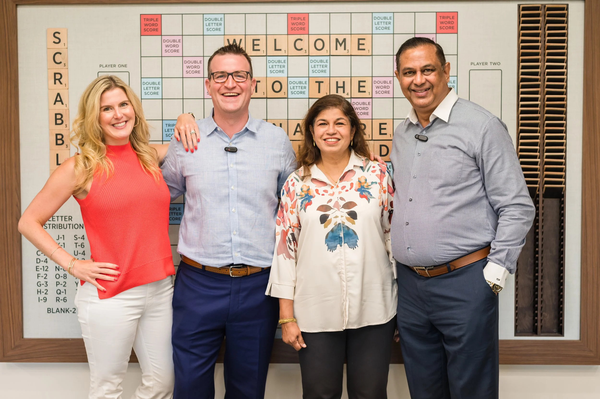 Four people smiling and standing together in front of a Scrabble board on a wall, with the words "WELCOME TO THE ADVANCED" spelled out on the board.