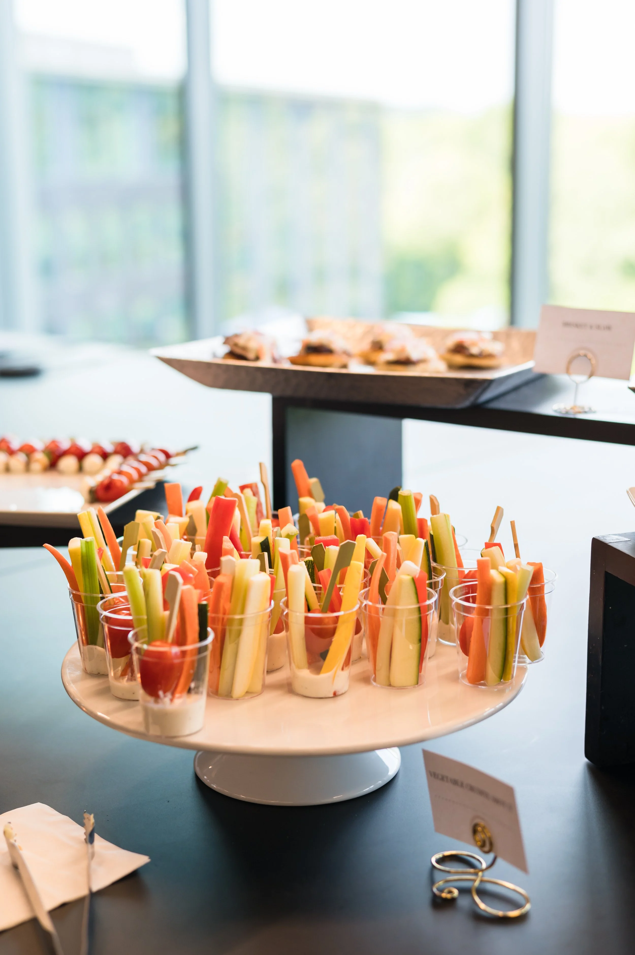 Assorted fresh vegetable sticks served in small cups on a white cake stand at a food buffet.