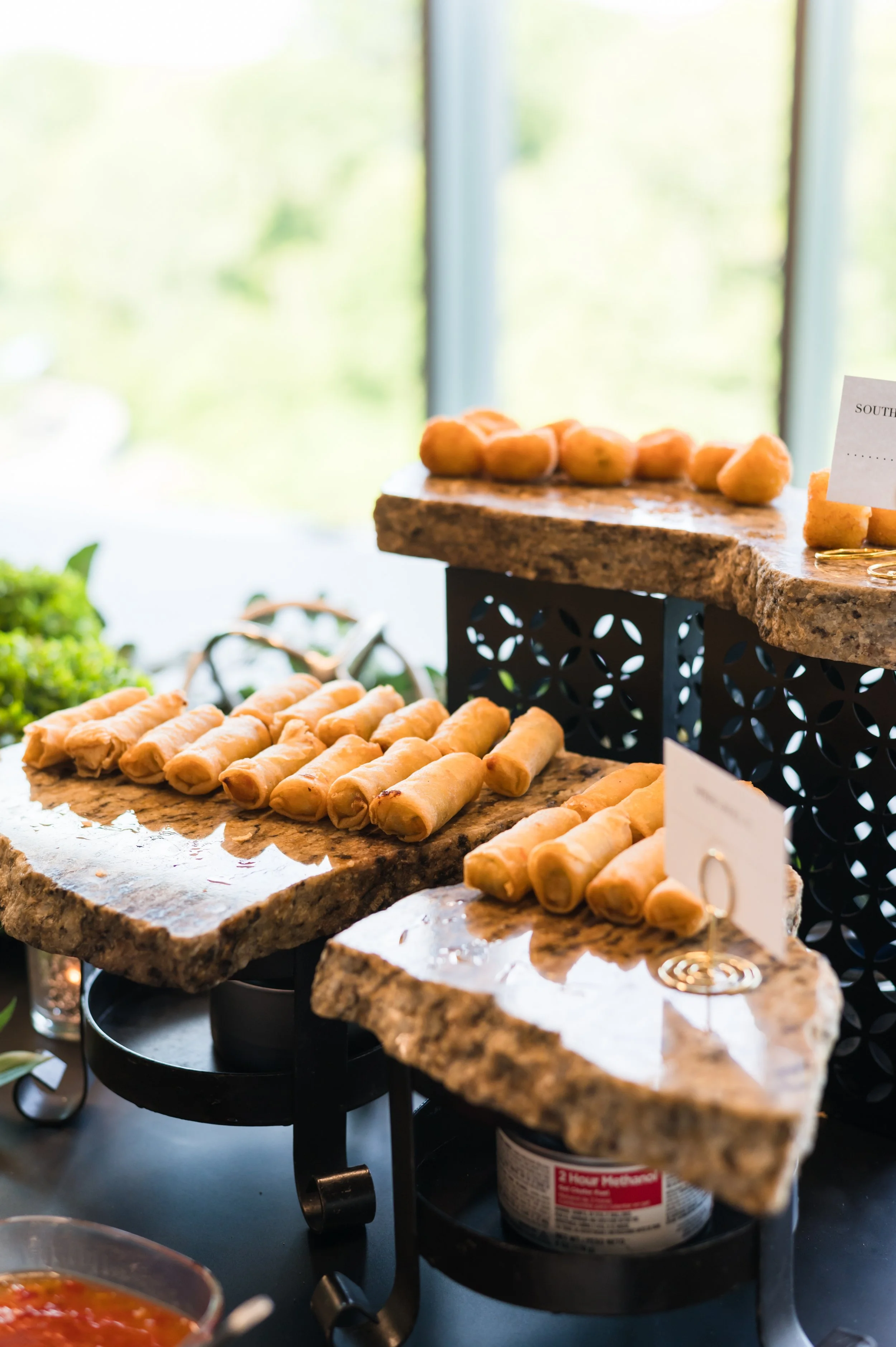 Display of neatly arranged spring rolls on marble slabs at a buffet or food station, with decorative greenery and a window in the background.