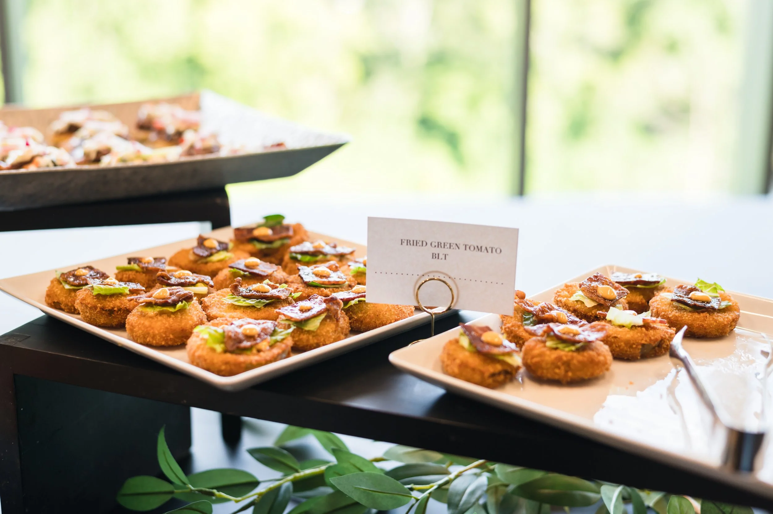 Buffet display with crispy fried green tomato bites topped with chocolate and nuts, labeled 'Fried Green Tomato BLT', on white plates.