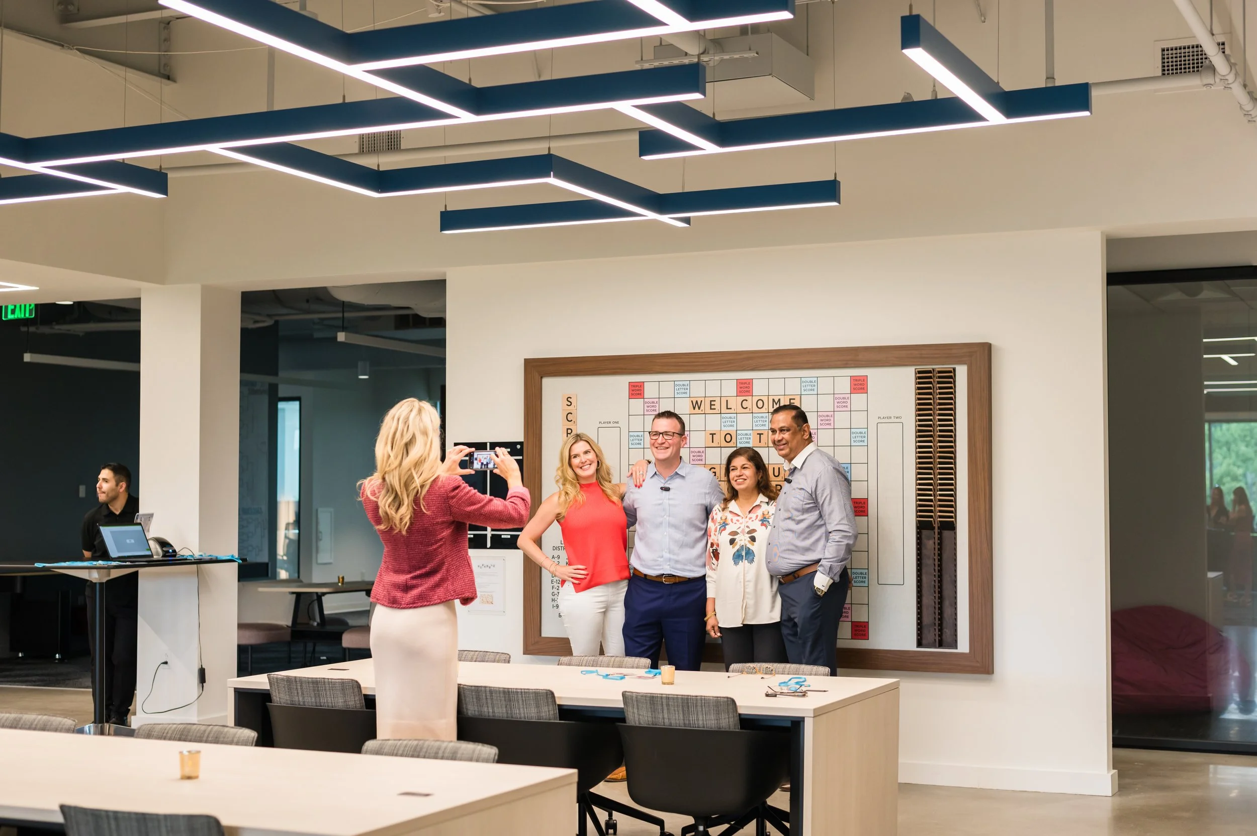 A group of four diverse adults standing together and smiling for a photo in an office setting. A woman in a pink blazer and light-colored skirt is taking the picture. A large scrabble-style welcome sign on the wall is behind them. The office has mode