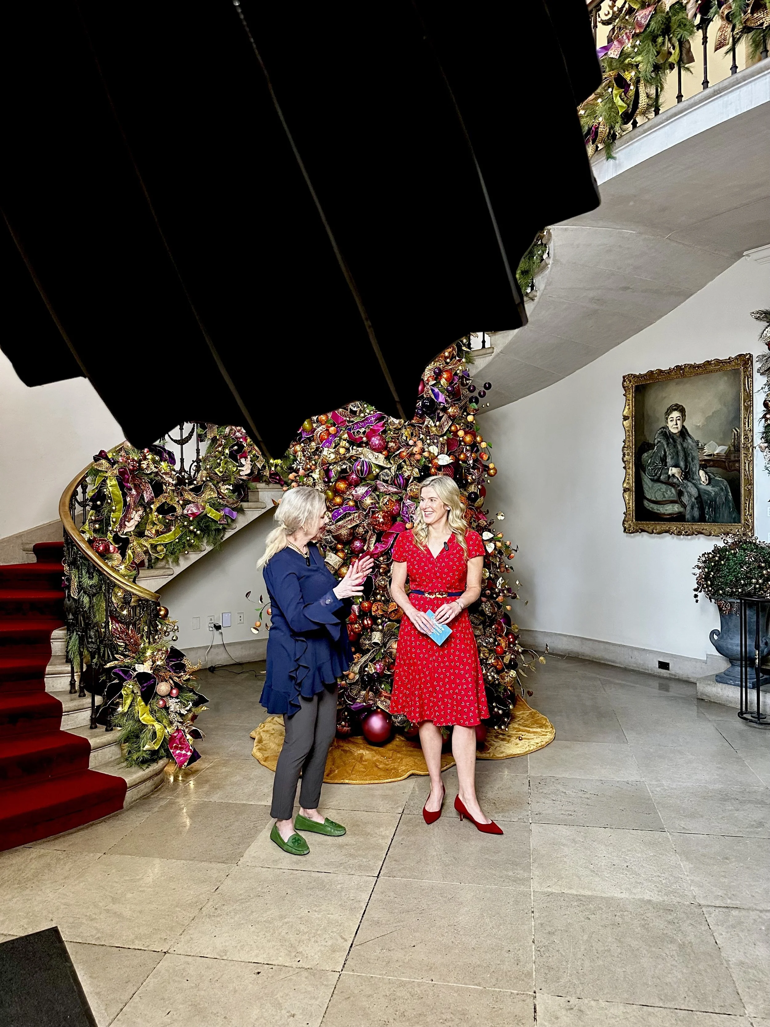 Two women conversing in front of a decorated Christmas tree indoors, with a portrait painting on the wall nearby.