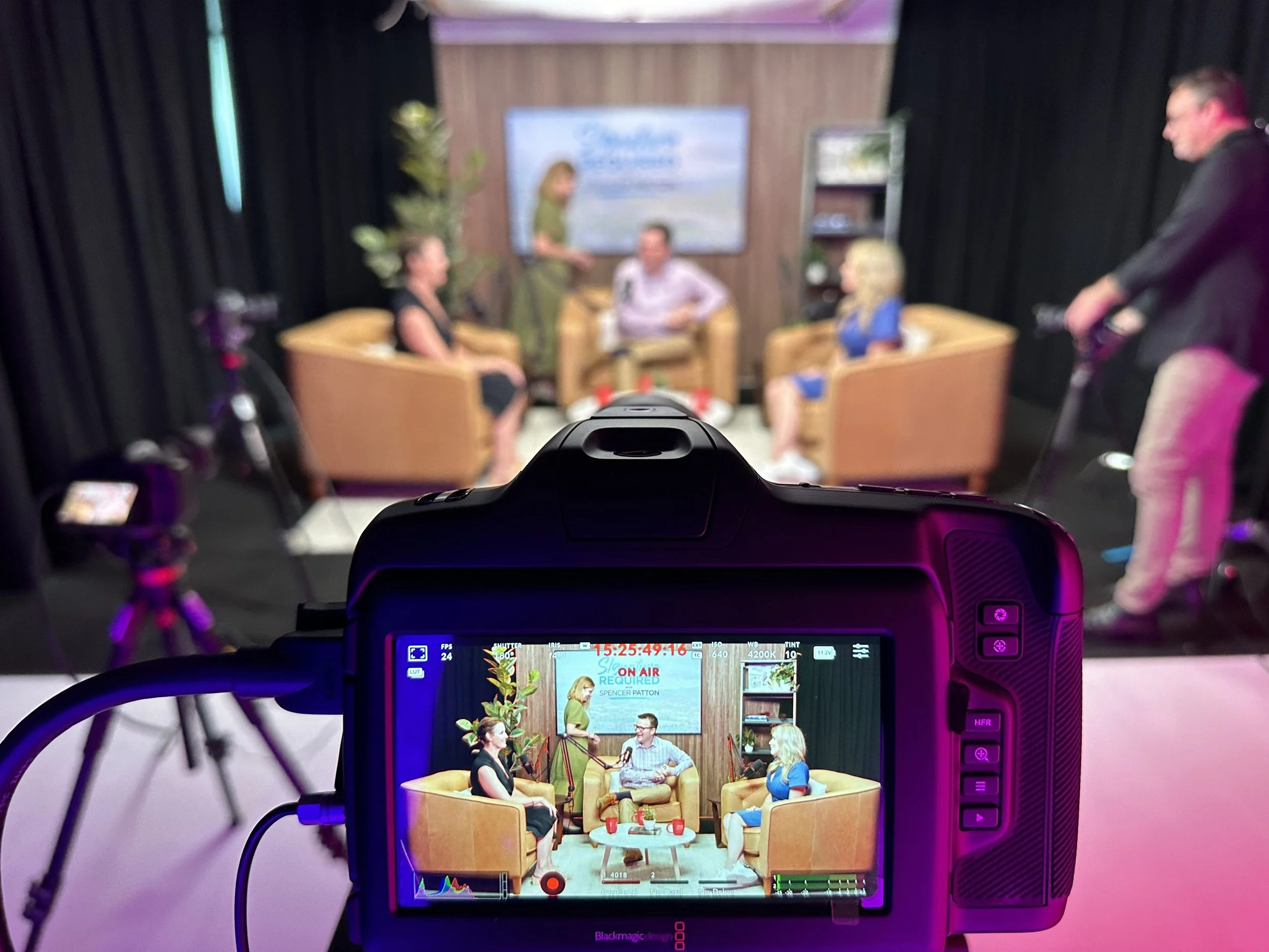 Television studio scene depicting four women and one man in a talk show setting. The camera view shows the scene on a monitor, with three women sitting in armchairs and a man in a chair. Two people are standing and preparing equipment, while a woman 