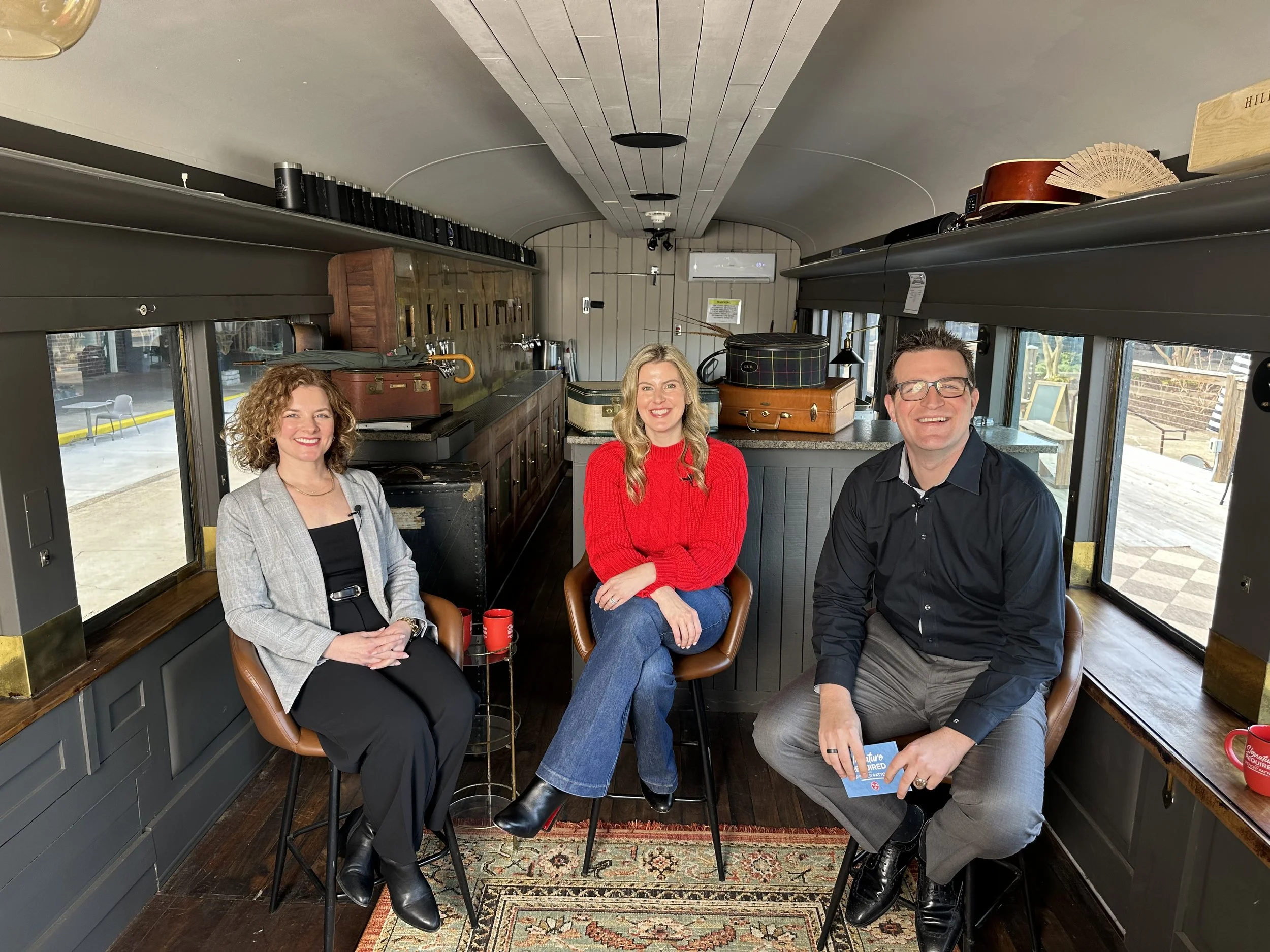 Three people sitting inside a vintage train car, smiling at the camera. Two women and one man, with the woman in the middle wearing a red sweater and the others in darker clothing. The train car has windows showing an outdoor seating area.