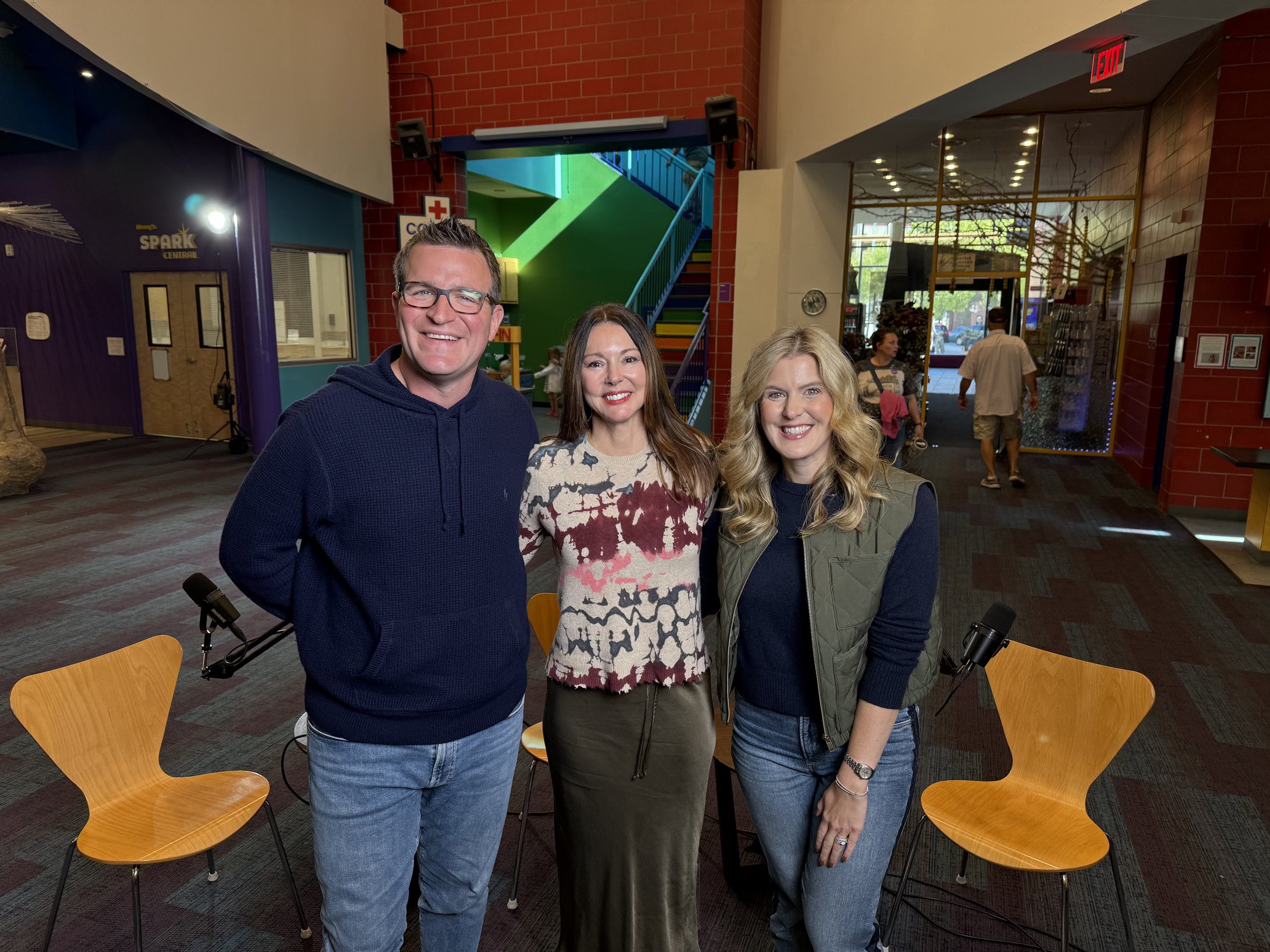 Three smiling adults, two women and one man, standing together inside a colorful indoor space with chairs, stairs, and people in the background.
