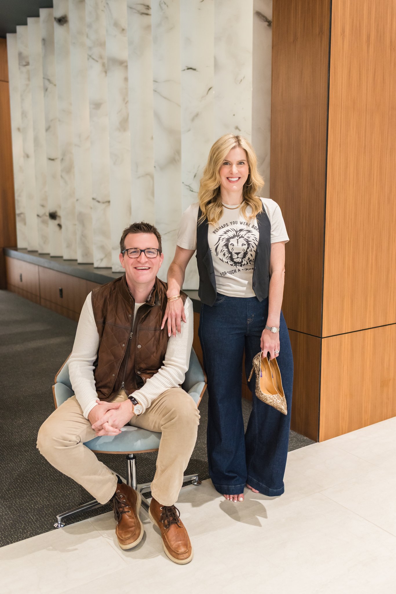 A man and woman posing together in an indoor setting. The man is sitting on a white chair, wearing beige pants, a white shirt, a brown vest, and glasses. The woman is standing beside him, smiling, wearing a white graphic t-shirt, a black vest, and wi