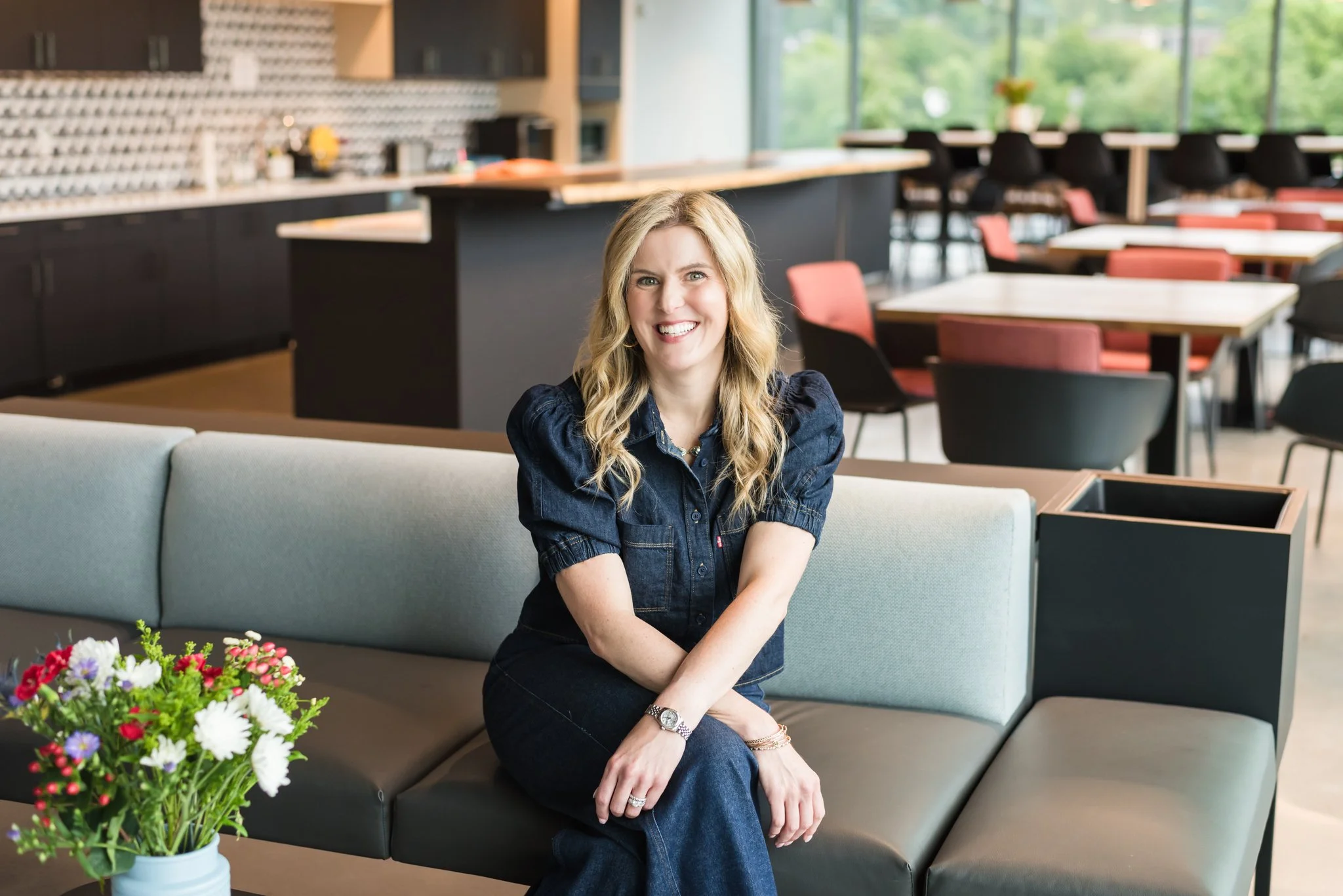 A woman with blonde hair smiling, sitting on a light-colored sofa in a modern dining area, with a vase of flowers on the table in front of her and an open kitchen in the background.