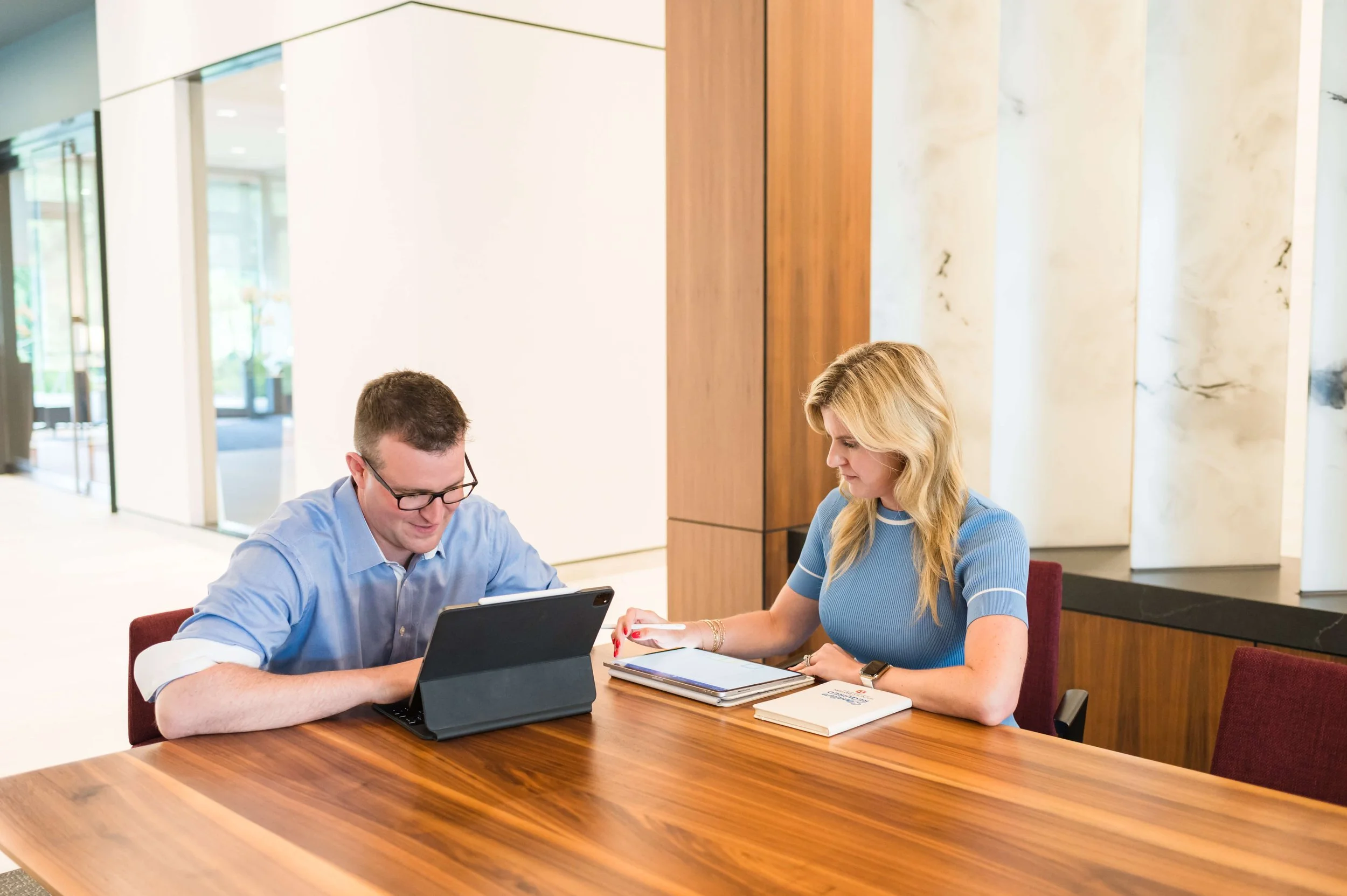 Two people, a man and a woman, sitting at a wooden table in a modern office or hotel lobby, looking at tablets or notebooks and engaged in a discussion.