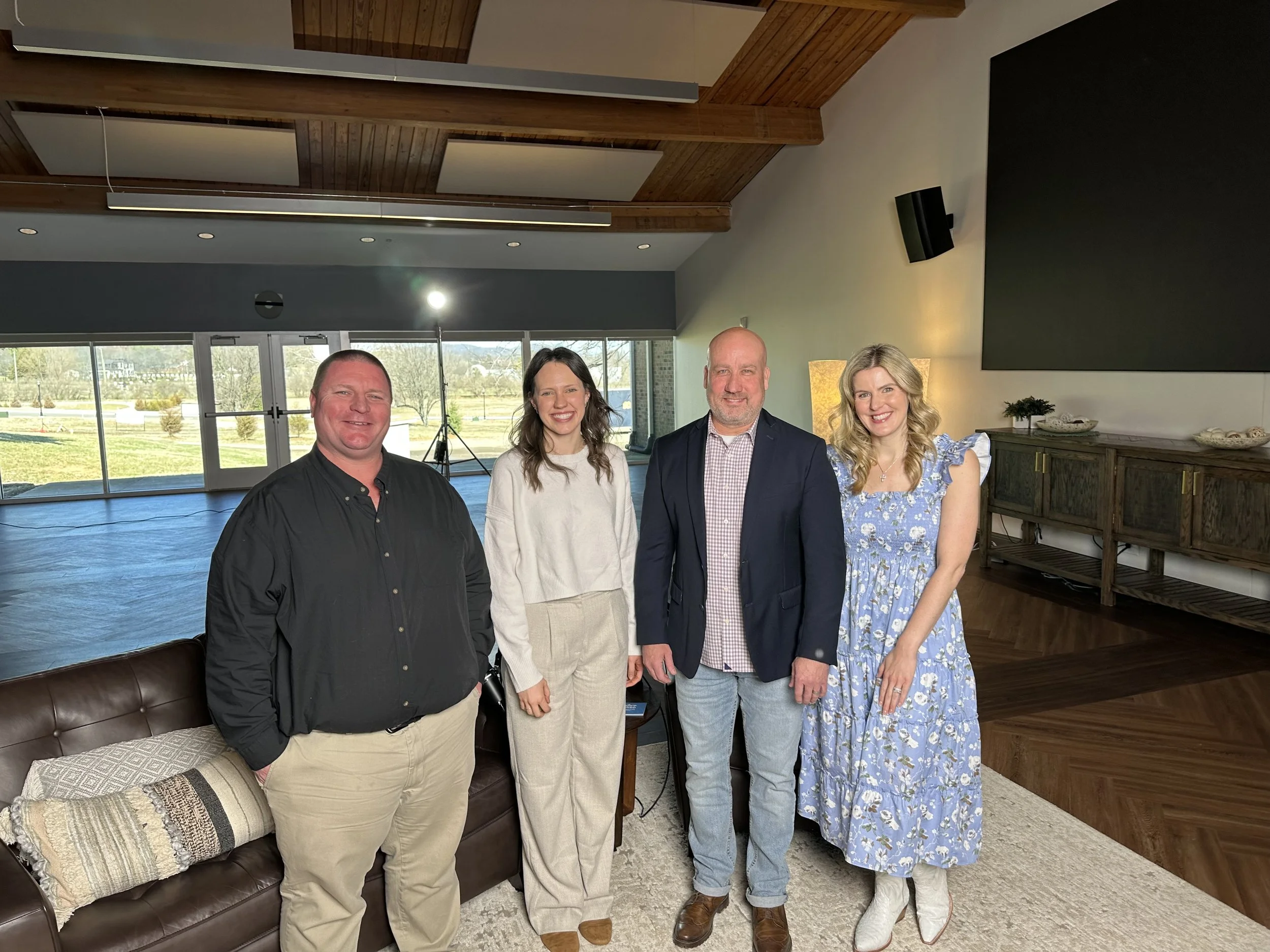 Four smiling people standing indoors in front of large windows that reveal an outdoor landscape with trees and a field, with a blue carpeted floor and a brown leather couch nearby.