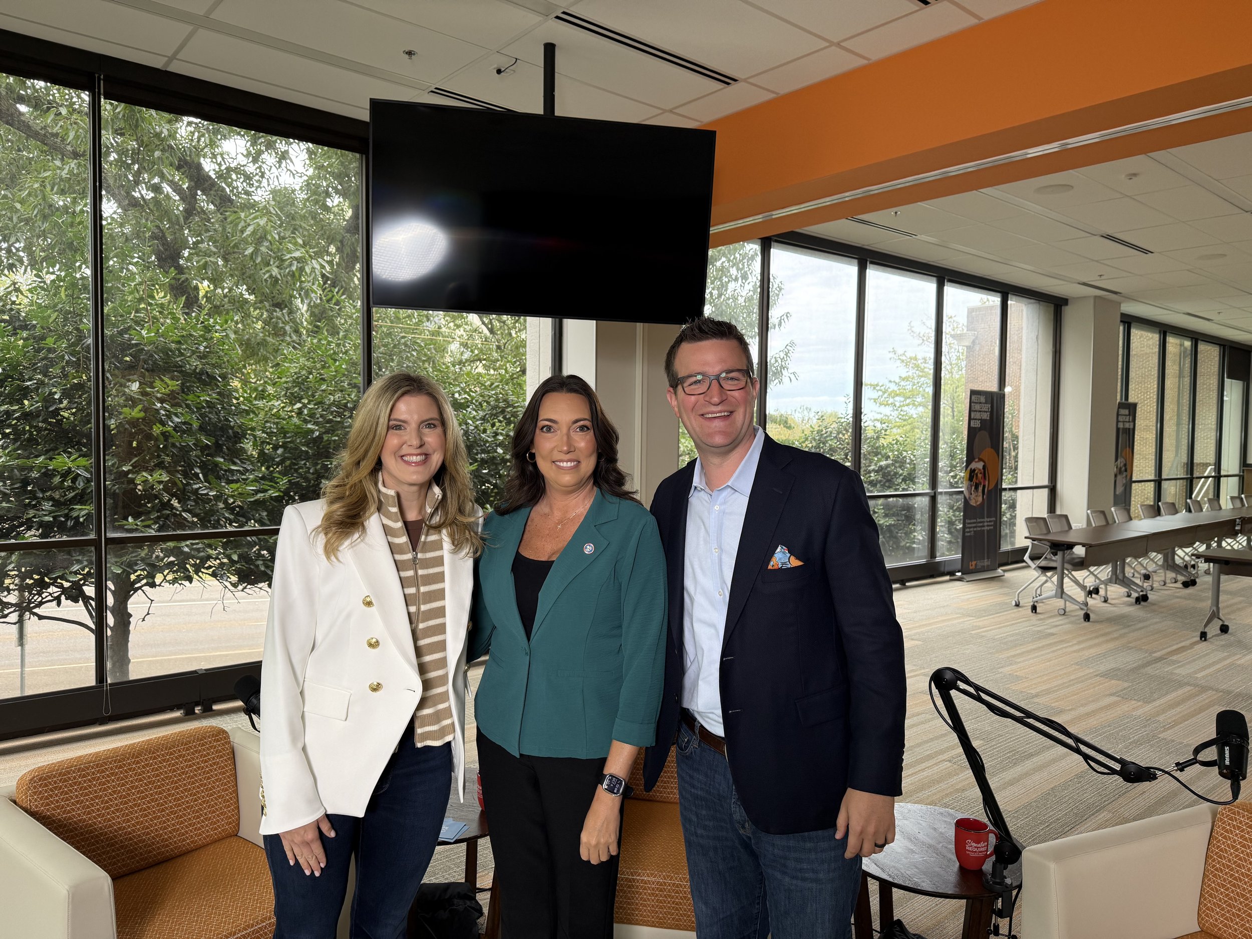 Three professionals standing in a modern office with large windows, posing and smiling for the camera.