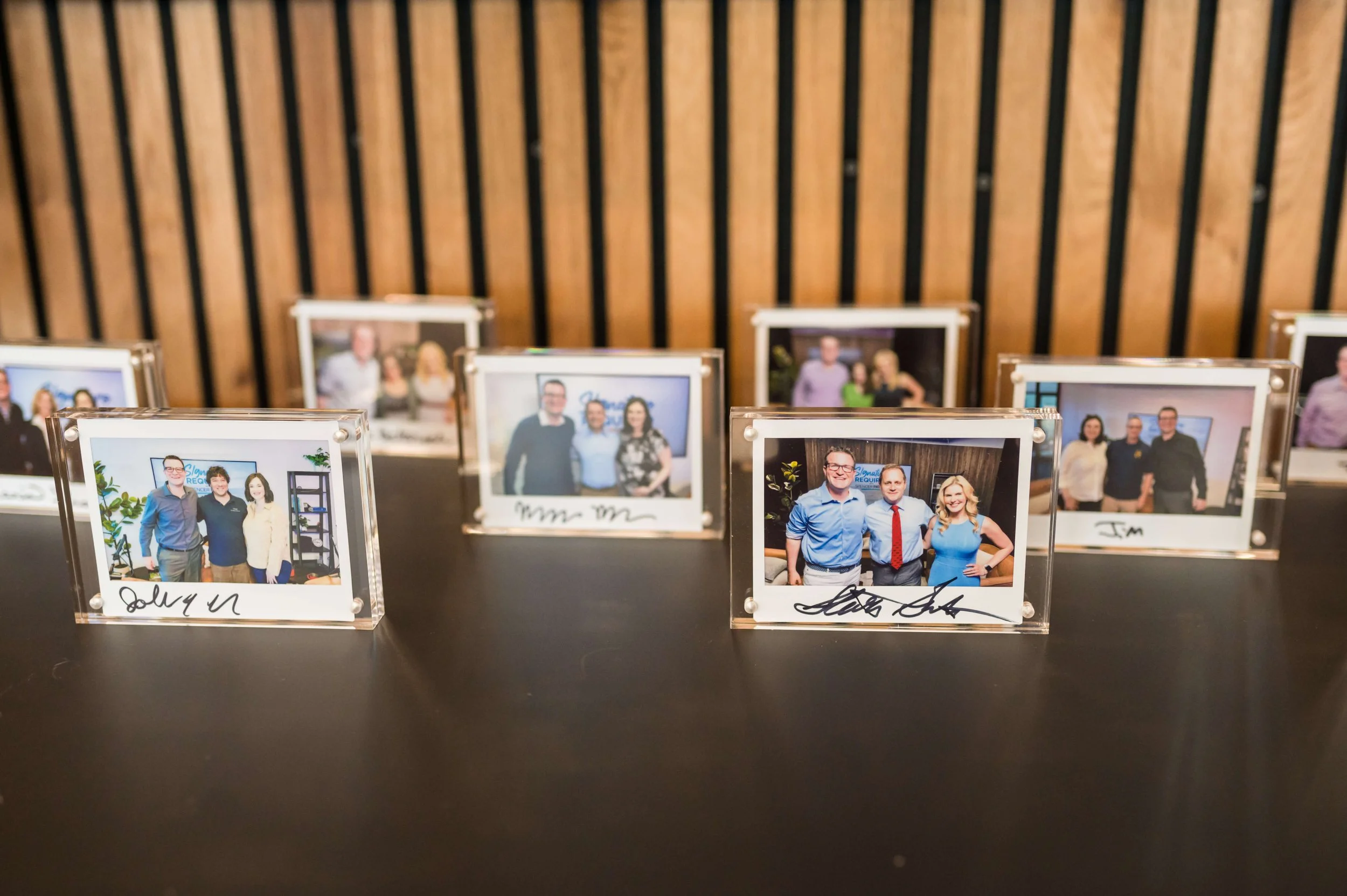 Photo booth with multiple framed pictures of people at an event, with signatures and dates written on the bottom of some frames, against a wood-paneled wall.