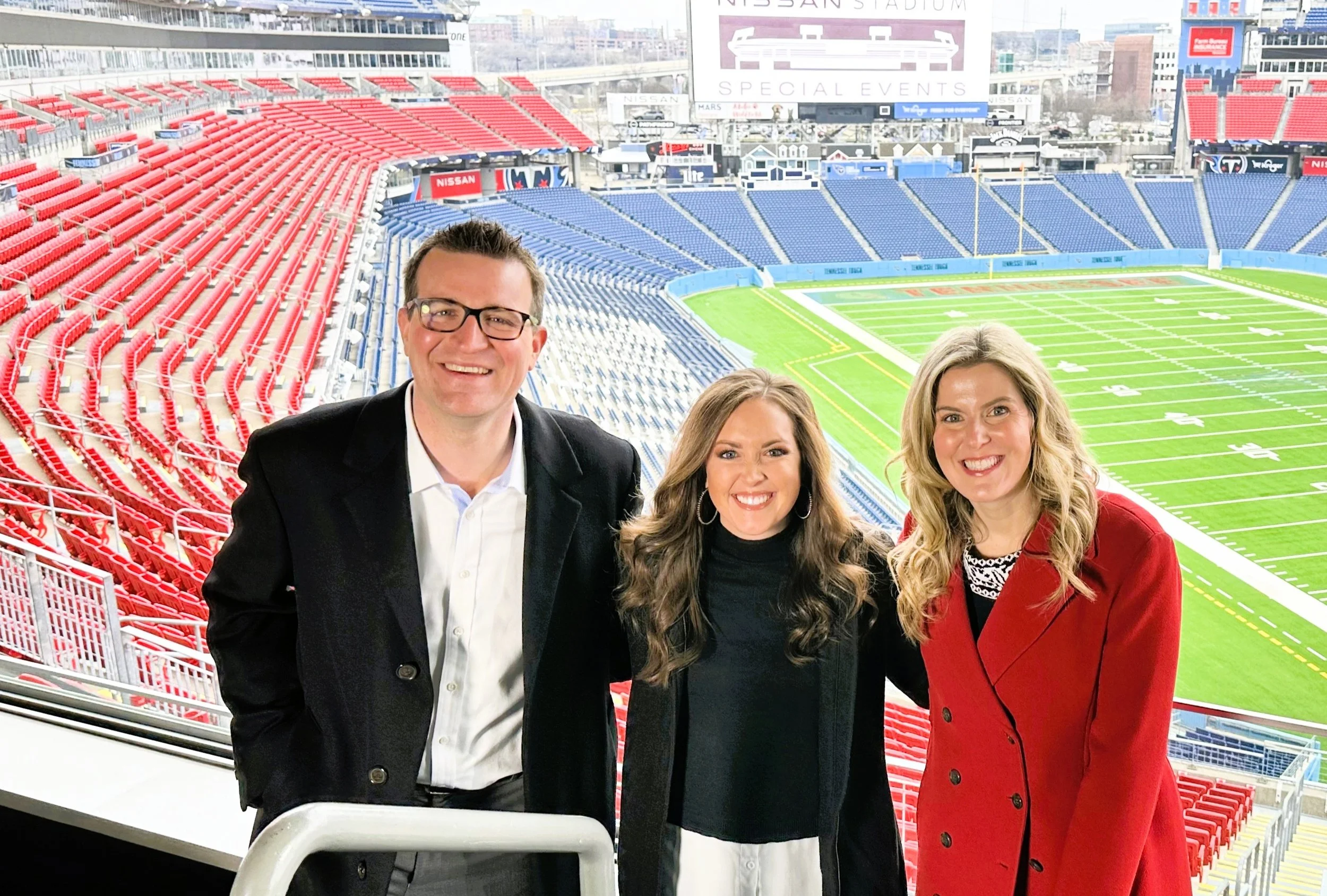 Three smiling people standing in a stadium with red and blue seats and a football field in the background.